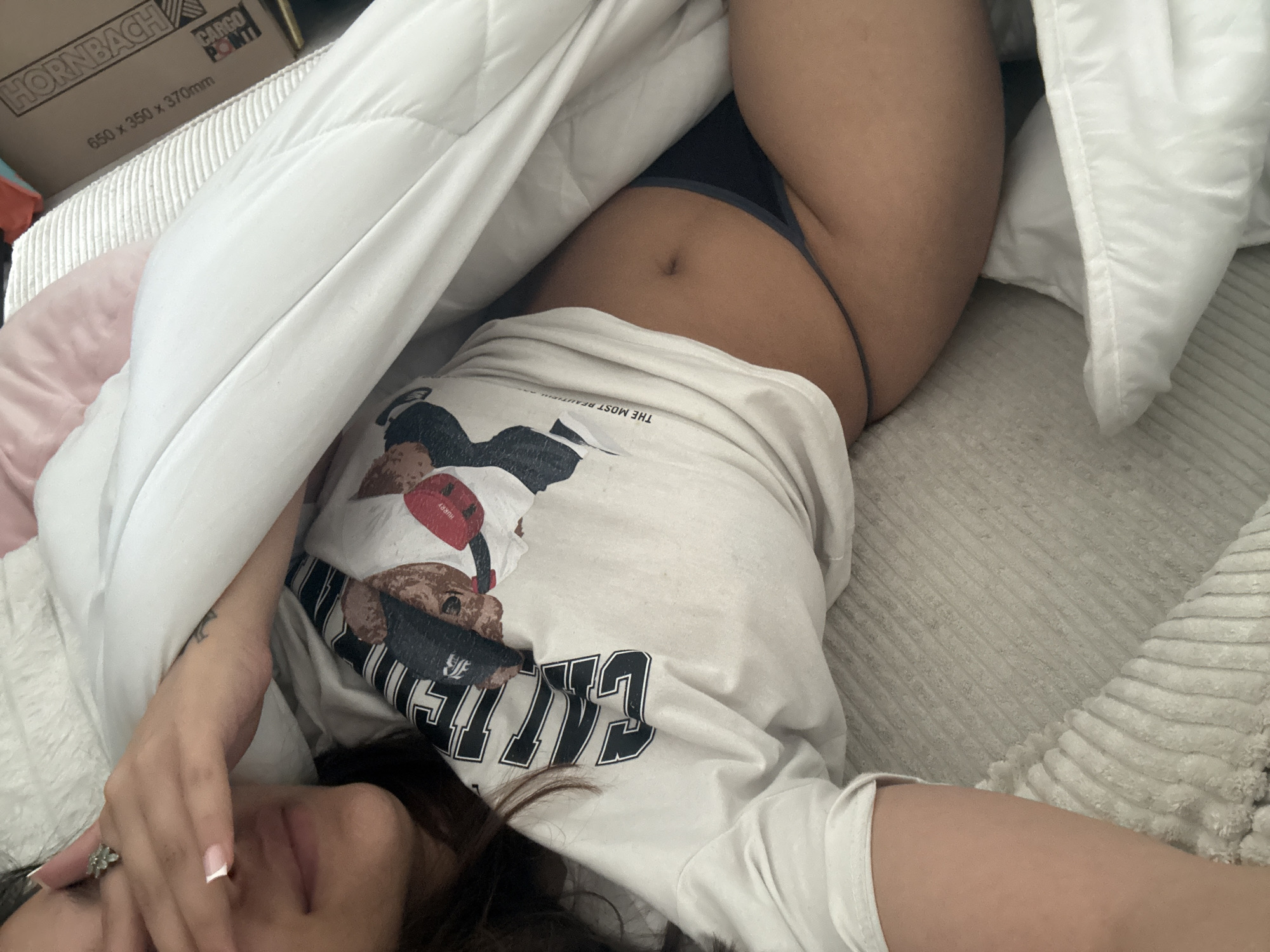 A woman is lying on a bed covered in a white comforter. She is wearing a white t-shirt and dark underwear. Her face is partially obscured by her hand. A cardboard box with the name "HORNBACH" and "CARGO POINT" printed on it is visible in the upper left corner of the image. The scene does not appear to be in the city of Mönchengladbach, Germany. The location cannot be determined.