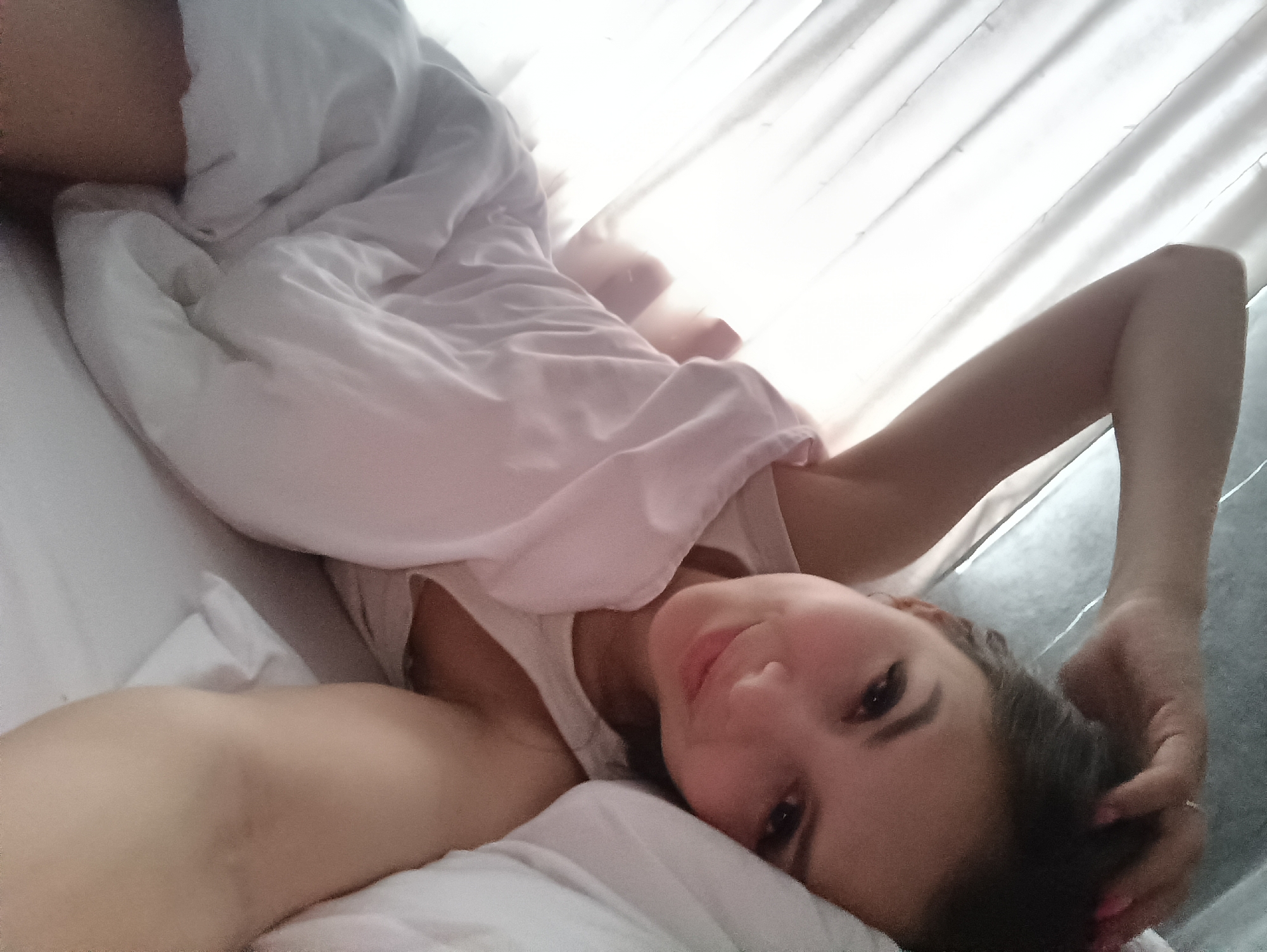 A woman is lying in a bed covered with white sheets. She is wearing a white tank top and has dark hair. Her arm is raised above her head, and her hand is touching her hair. The background shows a window with white curtains. The location is Unknown, Unknown.