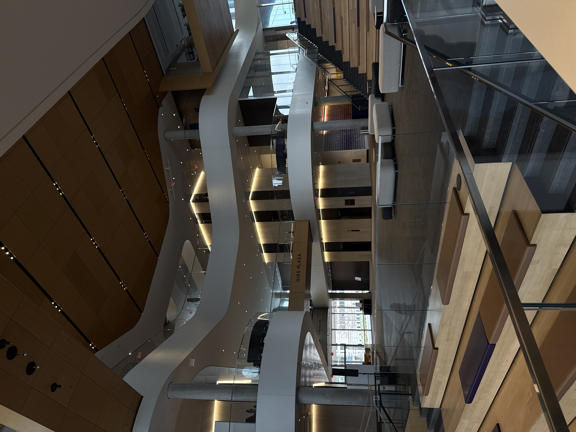 The image shows the interior of the Gies Business School at the University of Illinois Urbana-Champaign (UIUC), located in Champaign, Illinois, United States of America, not Evanston. The photograph is taken from a high angle, possibly from an upper floor looking down into the atrium.
The architectural design features curved white structures that appear to be support columns or walls, creating a modern and open space. There's extensive use of glass, allowing natural light to illuminate the area. The interior includes wooden paneling on the walls, which is arranged in linear patterns, contributing to the building's aesthetic.
The atrium includes seating areas with benches. Stairs and walkways are visible, suggesting multiple levels within the building. A sign for "GIES PLAZA" is present.
The environment seems to be an academic or professional setting, with an emphasis on contemporary design. There are no people in the view of the photo.