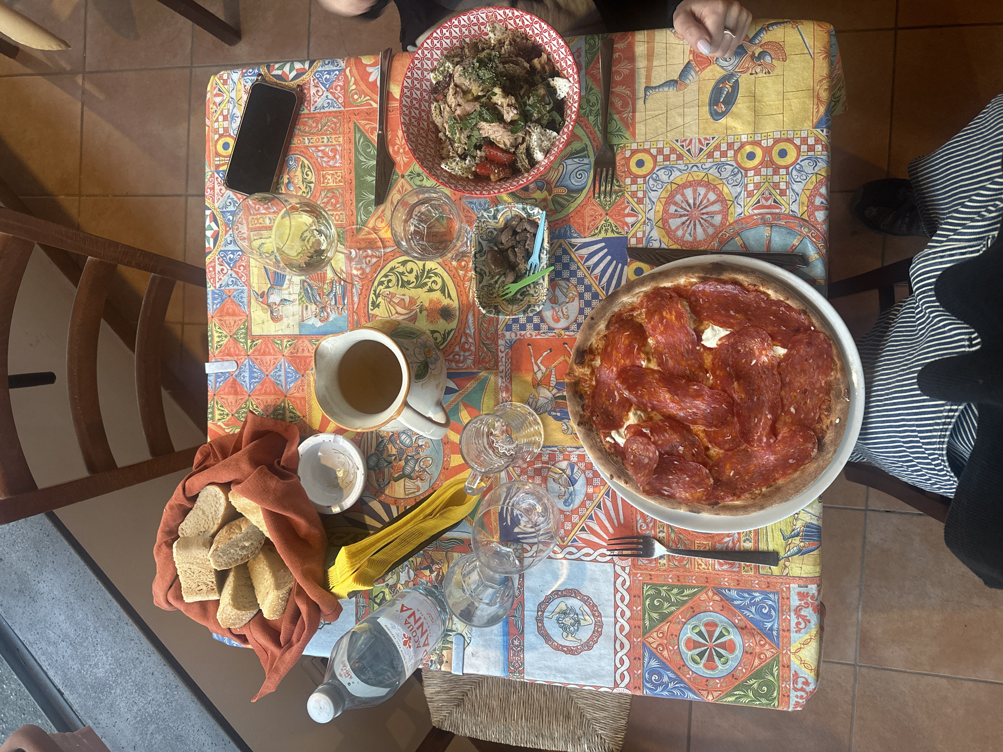 An overhead shot captures a dining table set for a meal, likely indoors at a restaurant or home. Two individuals are partially visible: one's hand with a ring and light blue nail polish rests on the tablecloth, while another's arm and striped shirt are seen on the right.
The table is covered with a vibrant, distinctly Sicilian-style patterned tablecloth, featuring geometric designs, colorful borders, and figurative elements such as triskelions (Trinacria symbols) and warrior-like figures in historical attire. This design strongly suggests a location in Southern Italy, aligning with the given context of Lipari.
Food and drinks are spread across the table. A large pizza, richly topped with pepperoni or salami and dollops of white cheese (possibly ricotta or fresh mozzarella), sits on a plate on the right. A generous bowl of salad containing tuna, leafy greens, tomatoes, and what appears to be crumbled cheese or another protein, is positioned at the top. A small, decorative dish holds olives and pieces of marinated meat or mushrooms, served with colorful plastic picks.
To the left, a rustic bread basket wrapped in an orange-brown cloth contains several pieces of crusty bread. Beverages include glasses of white wine, a pitcher filled with a light-colored liquid (possibly more wine or juice), and a plastic bottle of "ACQUA ANNA" branded mineral water, an Italian product. Several empty glasses, forks, knives, and yellow paper napkins complete the setting. A smartphone with a dark screen rests on the tablecloth near one of the wine glasses. The floor beneath the table is tiled in a warm, earthy tone.
The scene depicts a relaxed meal, likely during the daytime given the soft, ambient lighting. No specific emotions are discernible, but the arrangement suggests a pleasant social gathering.