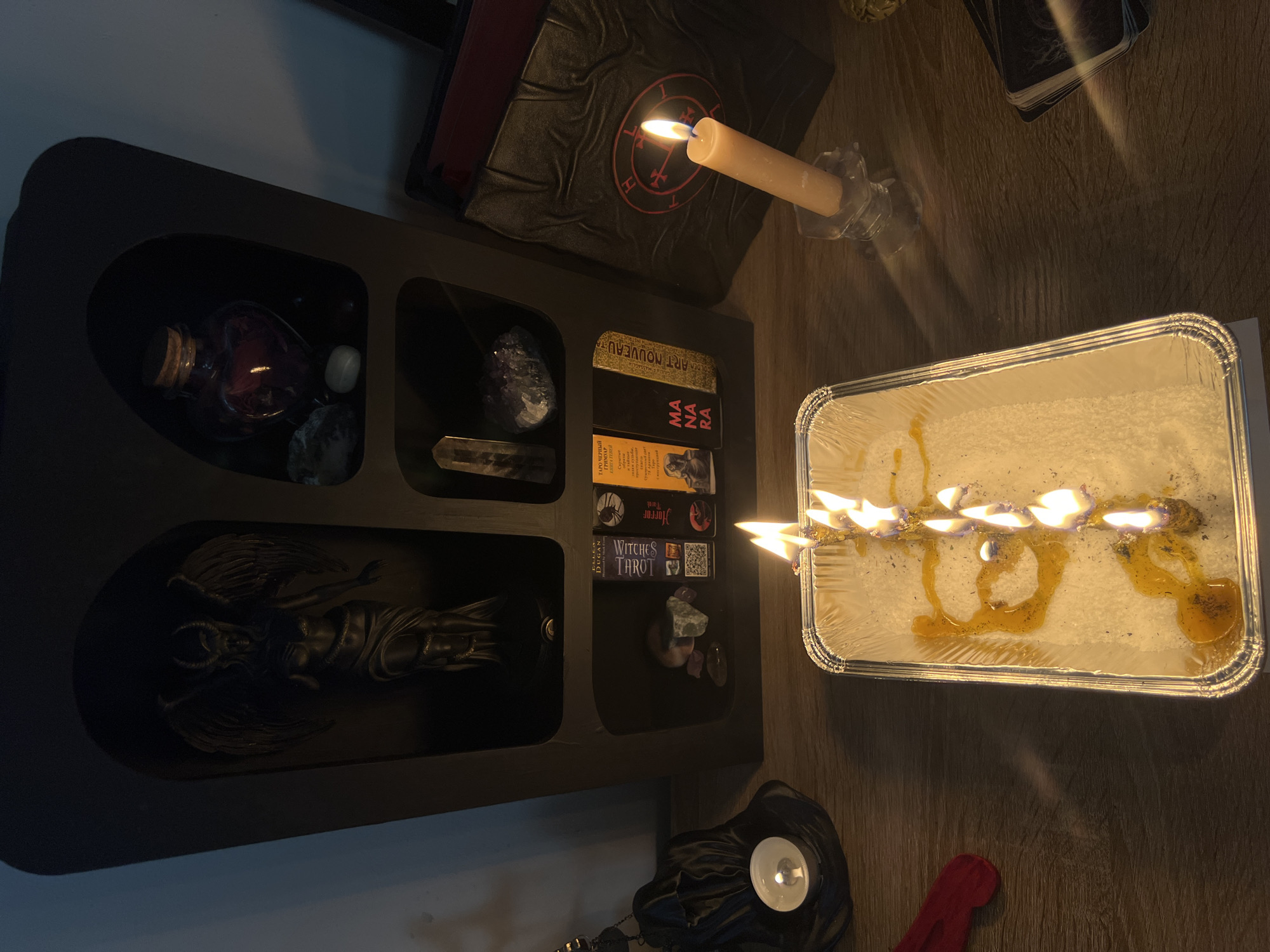 The image depicts a dimly lit, indoor scene with a collection of items that suggest a ritualistic or occult theme. In the upper left, a dark wooden shelf unit holds various objects. The top niche contains a dark bottle with a cork stopper, filled with a reddish liquid, along with some stones. The middle niche displays a large, dark crystal, a clear crystal, and a stack of tarot cards, including "Witches Tarot" and others with "MANA" visible. The bottom niche houses a dark, winged figurine, possibly Baphomet, with horns.
Above the shelf unit, a book with a circular, sigil-like emblem is visible, and a tall, white candle is burning, casting a warm glow. To the right of the shelf unit, a rectangular aluminum pan holds a white granular substance, possibly salt or wax, with several small flames burning along a horizontal wick or string. Yellowish liquid, likely melted wax or oil, is pooling in the pan.
In the lower right, a small, white votive candle in a black holder is partially visible, and a red cloth lies on the wooden surface. The overall mood is mystical and perhaps a bit unsettling due to the imagery and burning candles. The lighting is low, suggesting nighttime or a deliberately darkened room. No specific weather or location cues beyond the Polish context are evident.