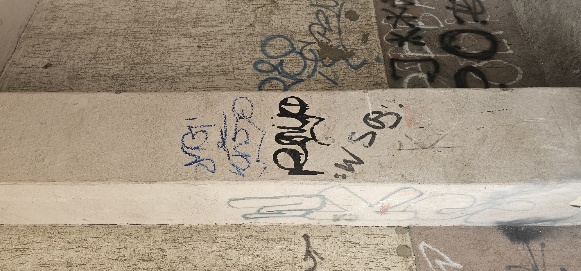 The image shows a close-up view of a concrete pillar covered in graffiti, located outdoors in Wrocław, Poland. The pillar is positioned at the corner of a building or structure, with two distinct textures of concrete visible on its surfaces. The left side of the pillar is textured with a rough, almost stucco-like finish, while the right side has a smoother, more even texture.
The graffiti is diverse in style and color. On the smoother, right-facing surface of the pillar, there are several tags and pieces. In blue spray paint, there are two distinct tags, one above the other, with the upper one appearing to be a stylized script. Below these blue tags, a large, black graffiti tag dominates the area, featuring bold lettering with drips. Below this black tag, there is another, smaller tag in black that reads "'ws g.". Further to the right, on the textured wall behind the pillar, there are more blue graffiti markings, appearing as interconnected loops and lines. There is also black graffiti on this textured surface, including what looks like asterisks and letters.
On the left-facing surface of the pillar, which has the rough texture, there are faint outlines of graffiti in light blue and white spray paint, including some vertical, elongated shapes. More graffiti is visible at the very bottom of the image, including black and white markings and what appears to be a darker, textured wall or ground surface.
The lighting suggests it is daytime, with shadows cast on the lower parts of the pillar, particularly on the right side. The overall impression is of an urban environment where graffiti is prevalent.