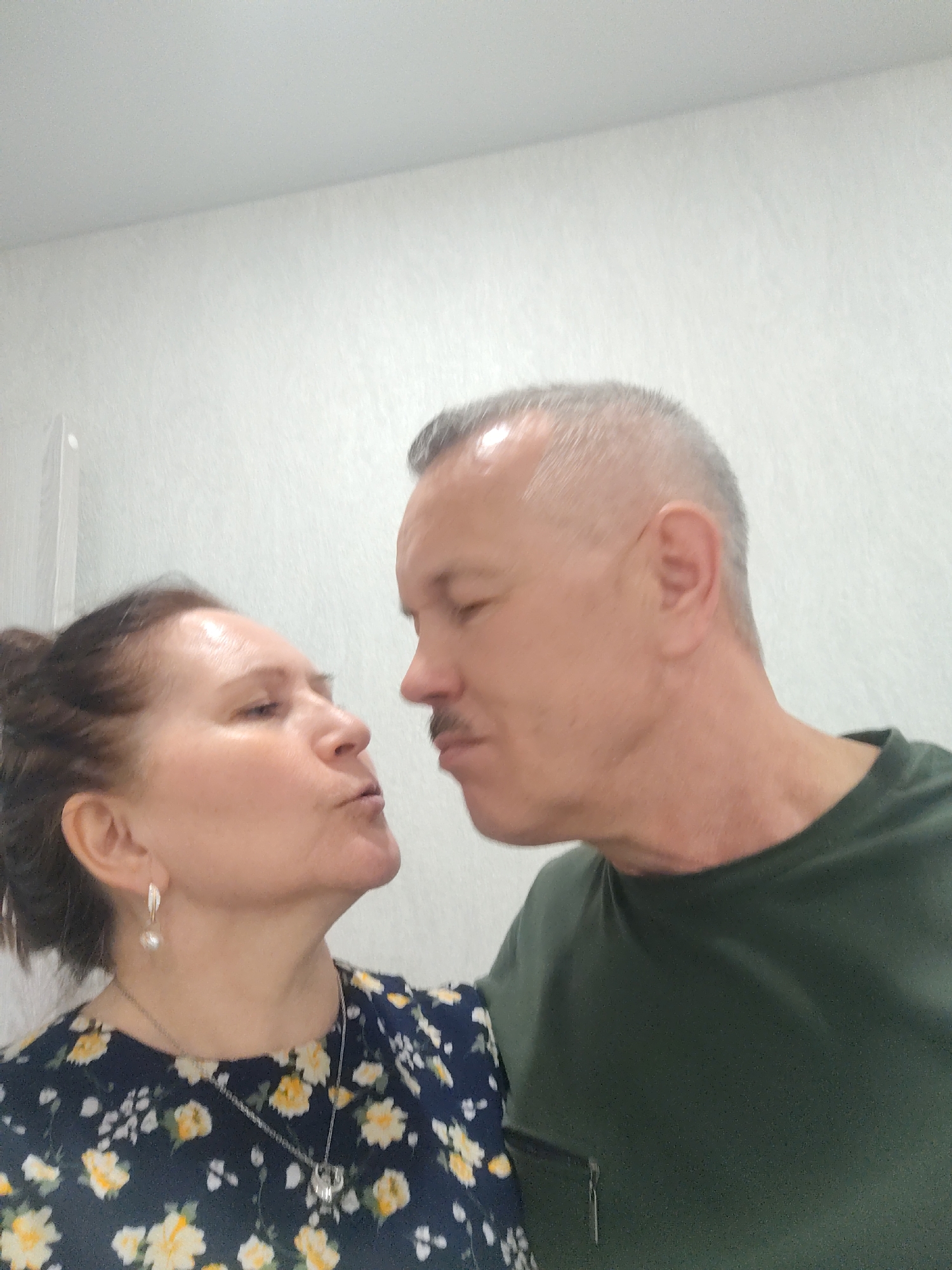 This close-up, eye-level indoor photo captures two middle-aged individuals, a woman and a man, in profile, facing each other with pursed lips, seemingly about to kiss or having just exchanged a kiss. The woman is on the left, looking up towards the man. She has dark hair styled up, wears pearl drop earrings, and a silver necklace with a pendant over a dark blue top with yellow and white floral patterns. The man, on the right, has short, graying hair, a light mustache, and is wearing a plain dark green crew-neck t-shirt. The background is a plain, light-colored wall with minimal texture and a faint horizontal line near the top. The overall mood conveys affection and intimacy. The image suggests a personal moment captured indoors, potentially in Sarapul, Russia, based on provided context. There is no visible text.