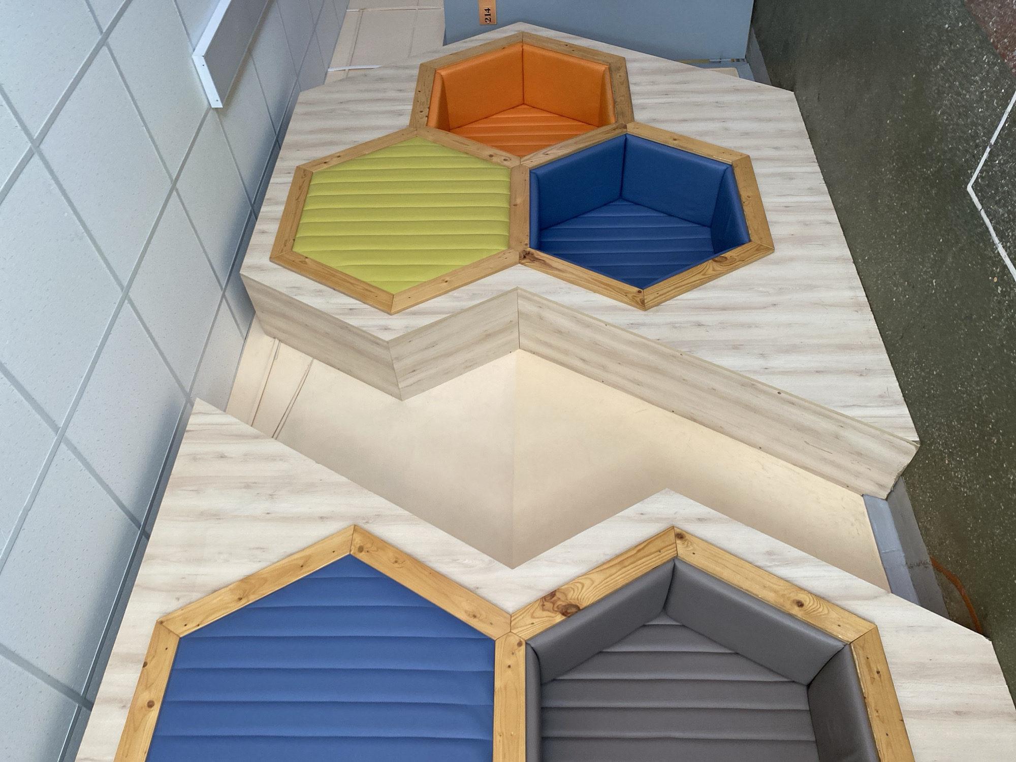 The image shows an elevated, modular seating area in a public space. The seating consists of several adjacent hexagonal or pentagonal units, each framed in light-colored wood and upholstered in a different color (orange, yellow, blue, and gray). The base and surrounding structure are finished in a wood-grain laminate. The location is determined as Votkinsk, Russia. The environment includes a light gray tiled wall on one side and a dark concrete floor on another, suggesting an indoor setting, possibly a library or community center. There are no people visible in the image. The scene is viewed from an elevated angle, looking down on the seating arrangement.