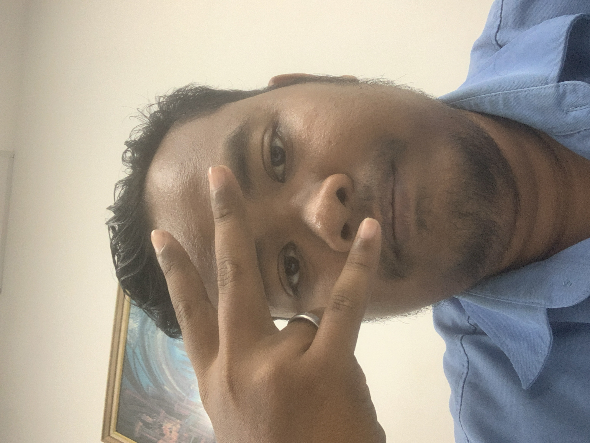 The image is a close-up selfie of a man with dark skin and curly black hair, wearing a blue collared shirt. He is making a peace sign with his right hand in front of his face, covering his left eye. He is also wearing a silver ring on his left ring finger. In the background, there is a framed painting with abstract art featuring blue and orange colors, and a plain off-white wall. The lighting suggests an indoor setting, possibly an office or a home. The photo appears to be taken from a chest-up perspective. There is no specific identifiable landmark to pinpoint the location in Palembang, Indonesia.