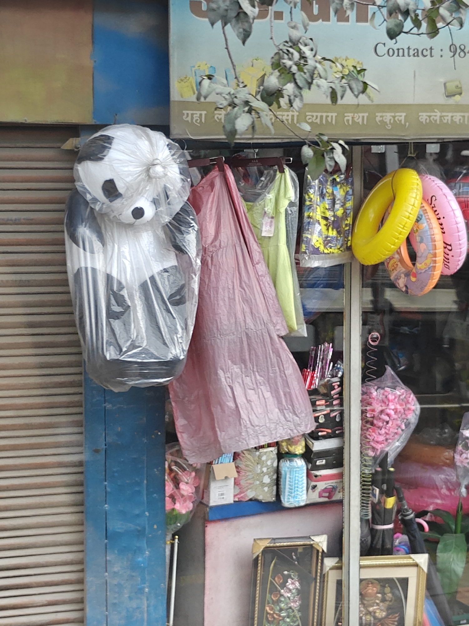 The image shows a storefront in Chandragiri, Nepal, displaying various items for sale. In the foreground, a large stuffed panda toy, wrapped in clear plastic, hangs from a metal rod. To the right, an assortment of items is visible, including a pink dress, several inflatable rings in yellow and pink, and other goods displayed on shelves within the shop. The shop's facade features a blue metal pillar on the left and a rolling metal shutter. A sign with Hindi text is visible above the shop window. The overall impression is one of a retail environment with a mix of toys, clothing, and other merchandise.