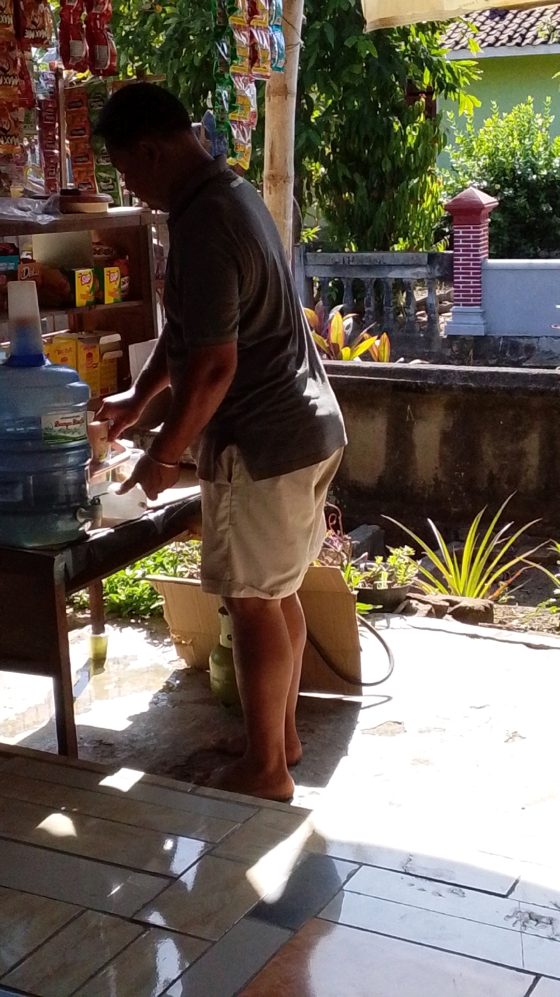 In a sunlit outdoor setting, likely in Lawu, Indonesia, a man is depicted dispensing liquid from a large blue water dispenser into a small cup. He is wearing a dark grey t-shirt and light beige shorts, and is barefoot. The dispenser is situated on a wooden table or stand, which also holds a few items, including what appears to be another water jug. In the background, various goods are displayed on shelves, suggesting a small shop or market stall. Beyond this, a textured concrete wall with a decorative railing and foliage can be seen, hinting at a residential or commercial area. The ground is a mix of concrete and tiled surfaces, with sunlight casting distinct shadows. A green gas cylinder is partially visible on the ground to the left.