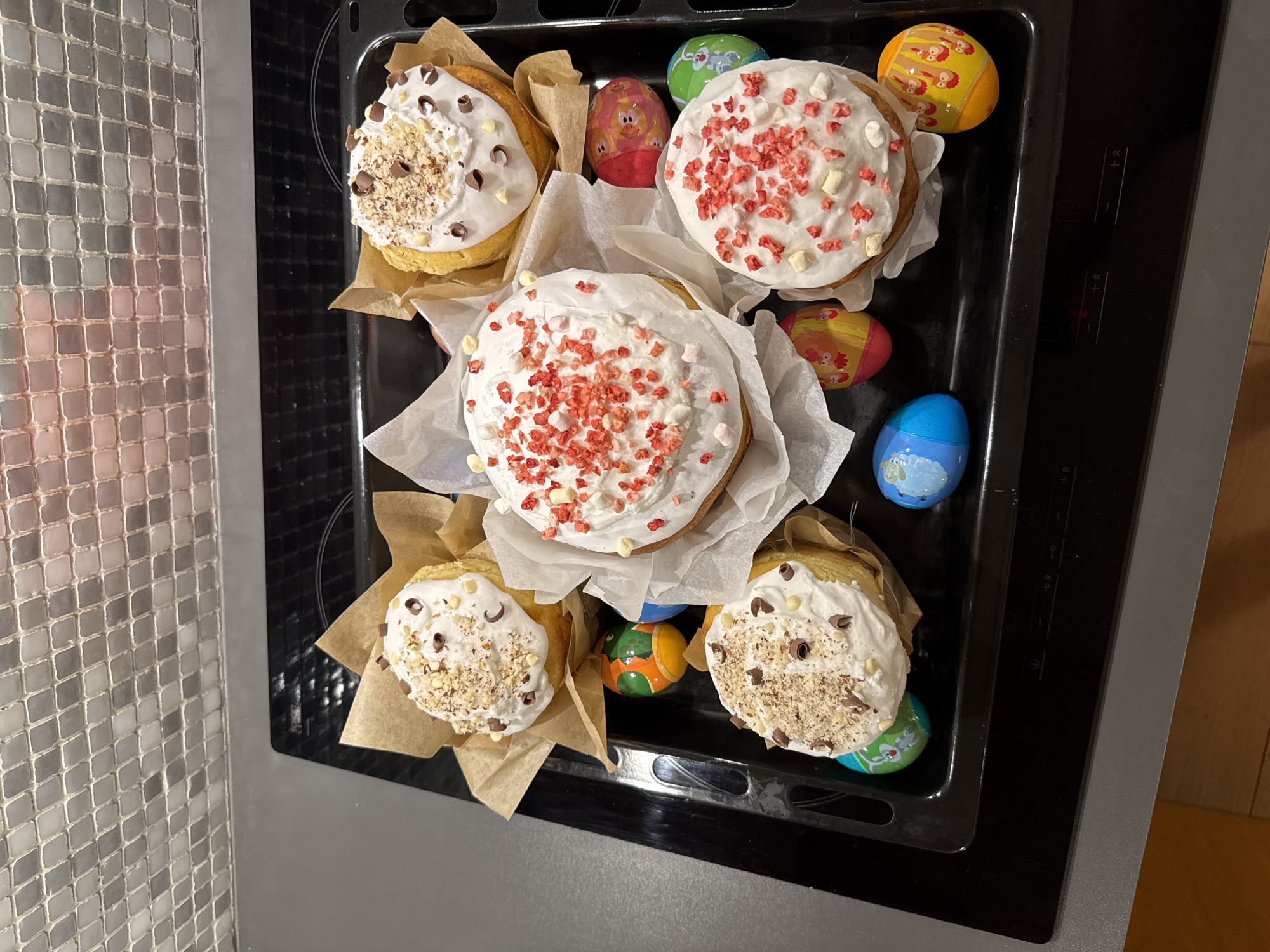 The image depicts a black oven tray containing five baked goods and several decorated Easter eggs. The baked goods appear to be Easter cakes, decorated with white icing and various toppings such as nuts, chocolate chips, and red sprinkles. The Easter eggs feature colorful designs with animal motifs. The tray is positioned on a grey countertop or surface next to a wall covered in small, square tiles. The location is likely a domestic kitchen. There is no evidence to identify Warsaw, Poland as the location.
