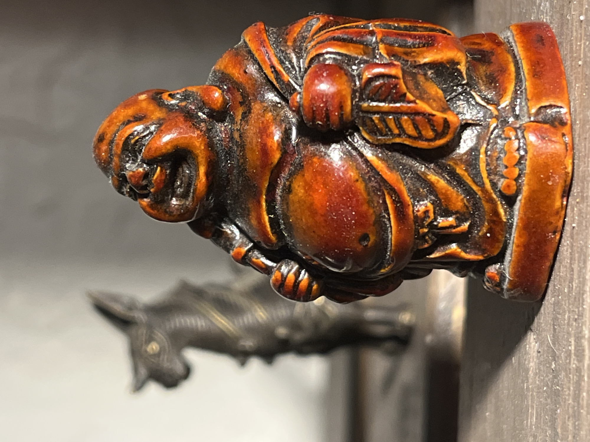 The image shows a small, ornate, dark brown or reddish-brown sculpture attached to a wooden surface. The sculpture appears to depict a figure with a somewhat grotesque or exaggerated face, resembling a lion or other animal with bared teeth. The body is bulky and detailed, possibly representing clothing or armor. In the blurred background, another sculpture, possibly of an animal such as a horse or donkey, can be seen. The setting is ambiguous, but seems to be indoors, possibly in a gallery, museum, or home. The specific location in Unknown, Unknown is not discernible from the image.