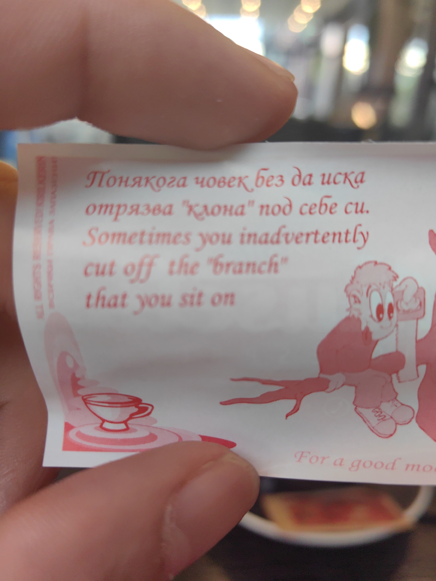 A close-up shot shows a fortune paper held by a person's hand. The paper has text written in Bulgarian and English: "Понякога човек без да иска отрязва 'клона' под себе си." and "Sometimes you inadvertently cut off the 'branch' that you sit on." Below the text, there is a cartoon illustration of a person sawing off the branch they are sitting on and an image of a teacup. The text "For a good mo..." is partially visible at the bottom. The location is likely in Sofia, Bulgaria, based on the Bulgarian text. The background is blurred, showing out-of-focus lights and a glimpse of a dish.