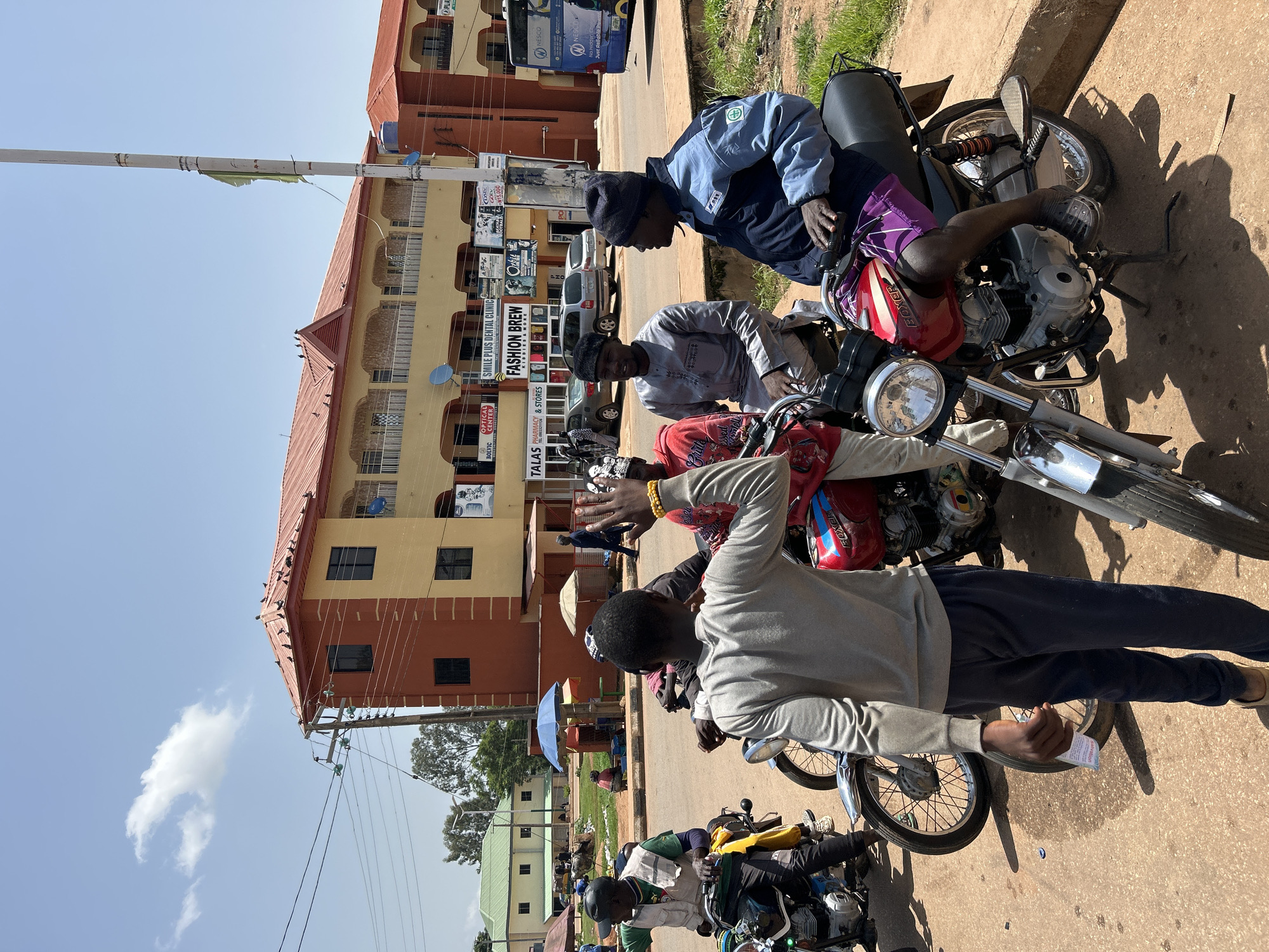 The image depicts a street scene in Bukuru, Nigeria. In the foreground, several men are gathered around motorcycles on a dirt road. One man in the center, facing away from the camera, wears a gray long-sleeved shirt and dark pants and holds a small white packet in his right hand. He appears to be interacting with another man to his left, who is wearing a green vest over a dark shirt and a black cap, seated on a motorcycle. To the right, two men are seated on a red motorcycle. One wears a blue jacket and purple shorts, while the other, wearing a gray traditional outfit and a black beanie, is looking to the right. In the background, a two-story building with orange and yellow facades stands prominently. Signs for businesses such as "FASHION BREW" and "SMILE PLUS DENTAL CLINIC" are visible on the building. A blue bus with "NESCO" branding is parked on the street. The environment is characterized by a clear blue sky with a single cloud, trees, and the presence of utility poles with electrical wires. The overall scene suggests a busy urban area.