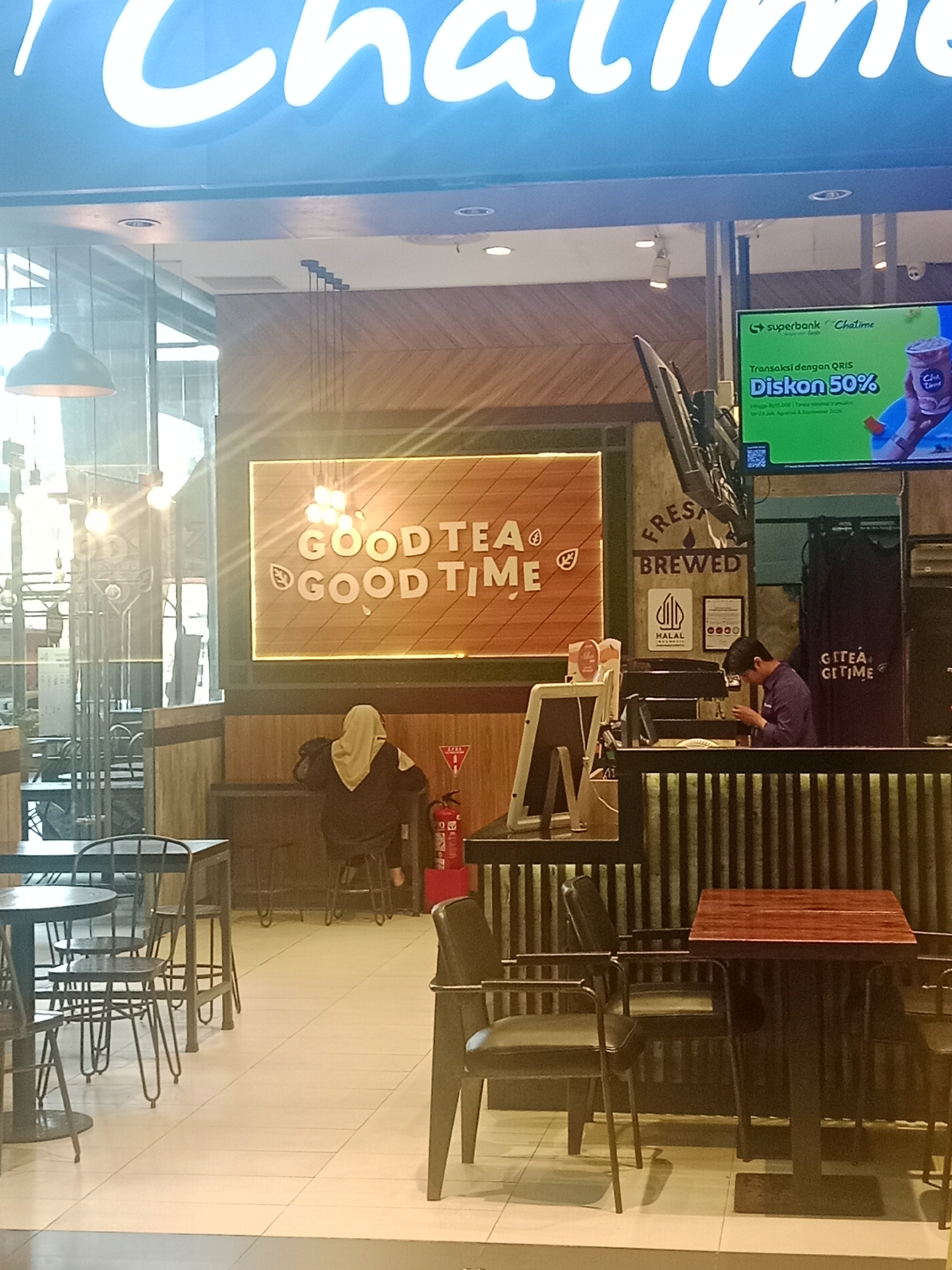The image depicts the interior of a Chatime outlet, likely located in Batam, Indonesia, given the context of its branding and the commonality of such establishments in the region. The scene is a casual dining/takeaway environment with multiple tables and chairs.
In the foreground, several dark metal tables and chairs are arranged, some circular and some square. The flooring is tiled in a light, neutral color. Behind these tables, a counter area is visible. A barista, dressed in a dark purple shirt, is attending to a customer or preparing an order behind the counter. Above the counter, there is a screen displaying an advertisement for a 50% discount on transactions made with QRIS, with the Superbank and Chatime logos prominent.
Further into the background, a woman wearing a hijab and a dark jacket is seated at a table, facing away from the camera. Behind her, a fire extinguisher is visible next to a wall. The walls of the outlet are decorated with wooden panels. A large illuminated sign with the Chatime logo is prominently displayed on the facade, above the entrance. The overall atmosphere suggests a busy but organized beverage shop.