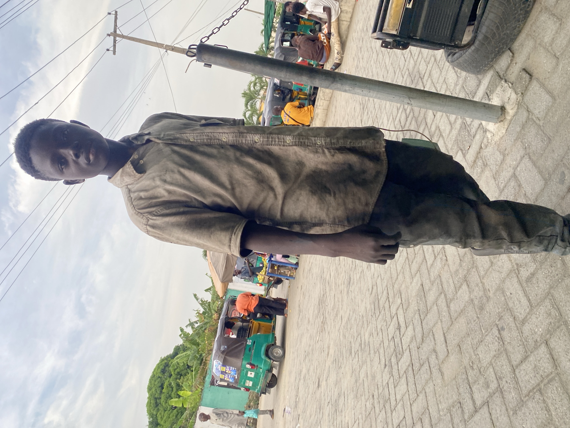 An outdoor, street-level scene in Lekki, Nigeria, captured under an overcast sky. The image features a young Black male in the foreground, facing the viewer with a neutral expression. He wears a faded, dark-colored long-sleeved button-up shirt and dark pants.
The ground is paved with grey interlocking bricks laid in a herringbone pattern. A sturdy metal utility pole stands prominently in the mid-ground, extending vertically. Power lines are visible in the upper left section of the sky. To the right of the main subject and behind the pole, a bustling street environment unfolds. Several people are visible, including one wearing a bright yellow shirt and another in an orange shirt. Multiple green auto-rickshaws, commonly known as "keke napep" in Nigeria, are parked or moving along the street. One such keke has the number "05" faintly visible on its side. In the background, dense green foliage, consistent with tropical trees like banana plants, lines the area. On the far right, part of a black generator or similar equipment with a tire is visible.
The lighting is soft and diffuse, indicating an overcast day. The main subject appears to be posing for the camera, while people in the background seem to be going about daily activities, likely involving public transport or commerce.