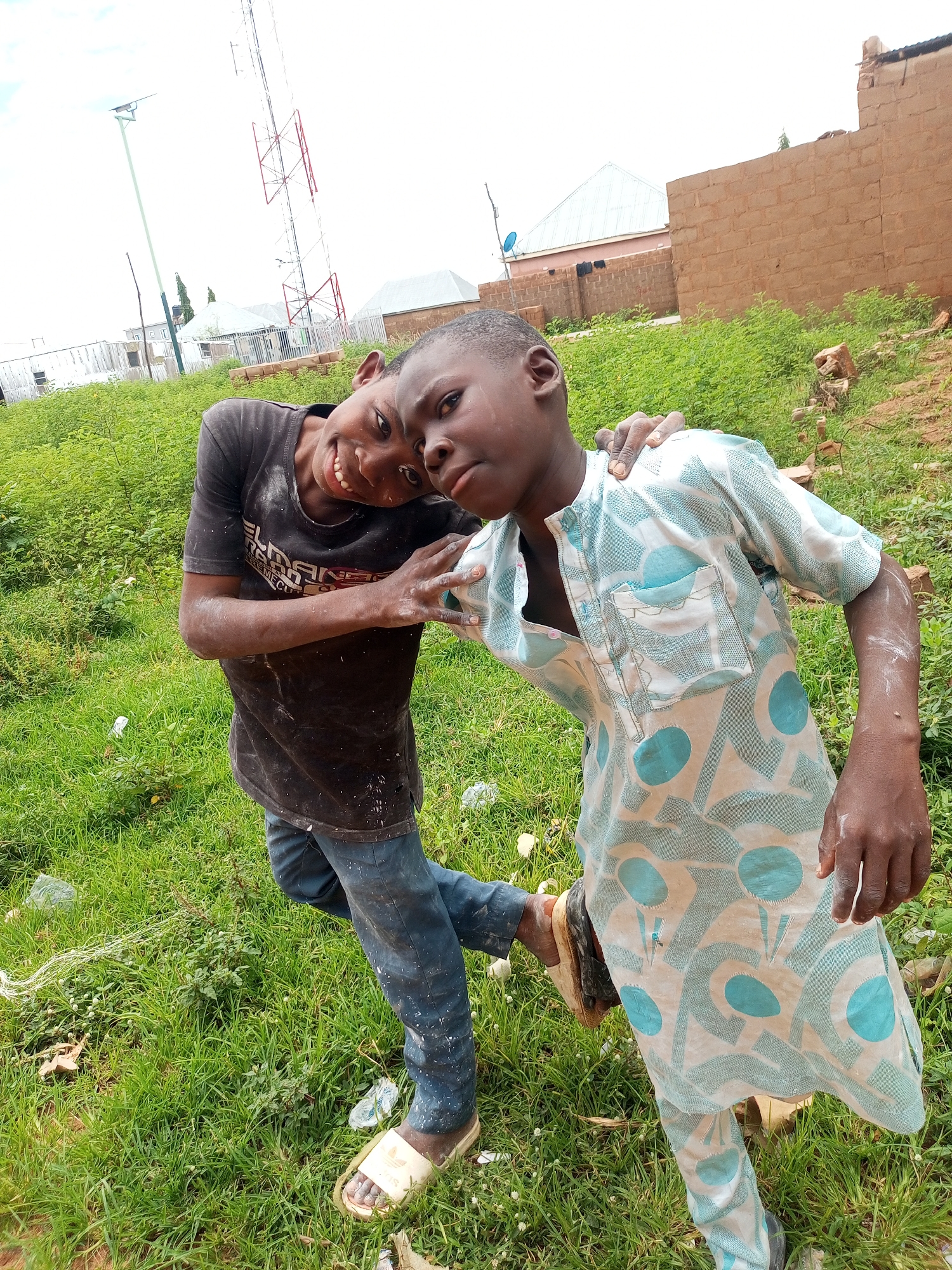 Two young boys are posing for a photo outdoors in what appears to be a semi-urban area of Tempure, Nigeria. The boy on the left is wearing a dark t-shirt with white splatters and dark pants, and is leaning his head on the shoulder of the boy on the right. He is smiling broadly and has his arm around the other boy's shoulders. The boy on the right is wearing a light-colored, patterned traditional African garment and dark pants. He is looking towards the camera with a neutral expression. They are standing on a grassy area with some small white flowers. In the background, there is a tall radio tower, a solar-powered street light, some brick buildings, and dense green vegetation. The ground is uneven and appears to be a mix of dirt and grass. There is some scattered litter on the grass. The sky is overcast and bright.
