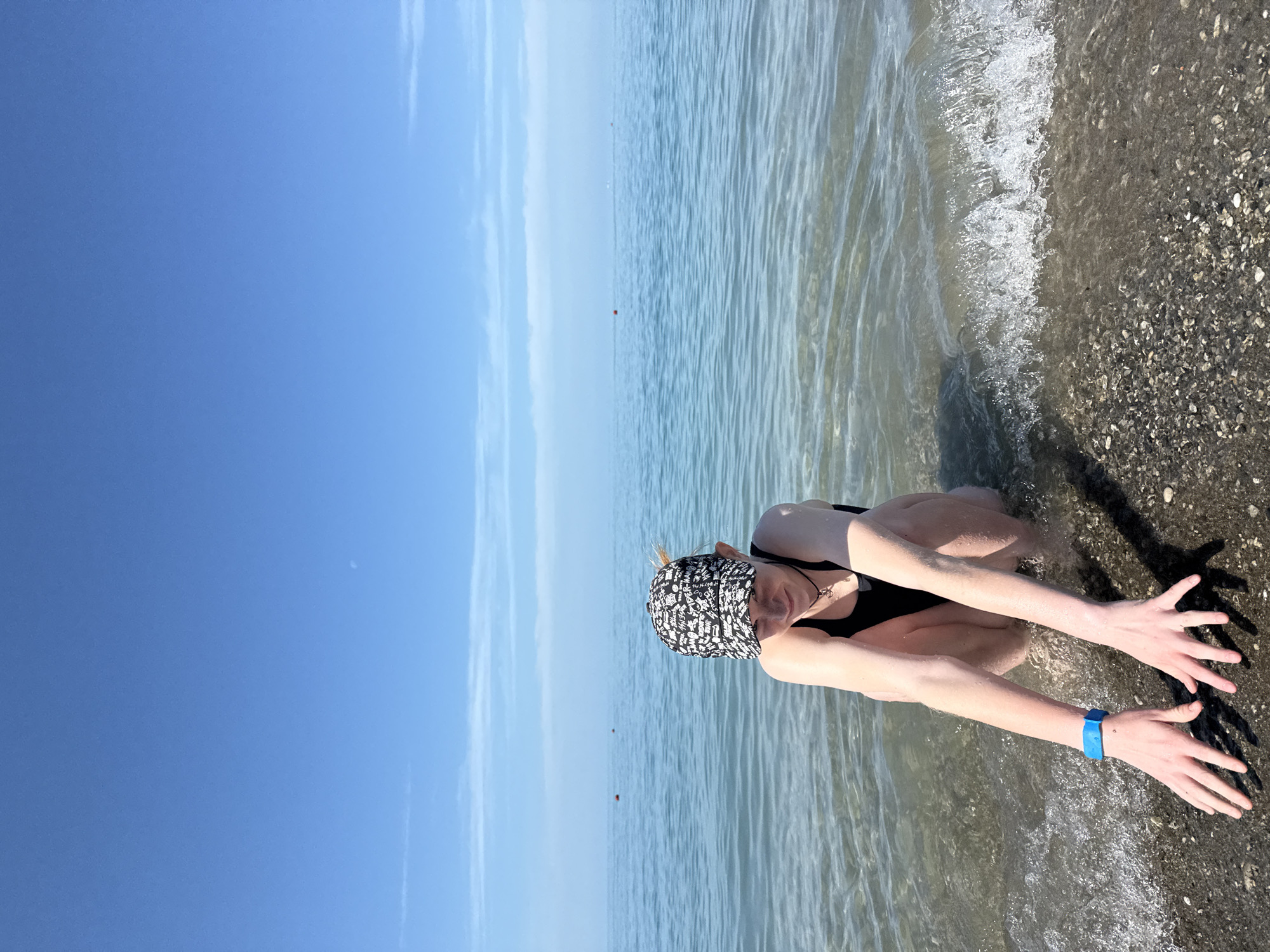 The image depicts a young person crouching at the edge of the Black Sea on a pebble beach in Nebug, Russia, under a clear blue sky.
A light-skinned individual, appearing to be a young female, is positioned centrally in the lower half of the frame. She wears a black one-piece swimsuit, a black-and-white patterned baseball cap featuring "Spider-Man" motifs, a dark choker-style necklace with a small metallic pendant, and a bright blue wristband on her left wrist. Her head is tilted down, and her gaze is directed towards her extended arms. Both hands are resting in the shallow, clear water and wet, dark pebbles at the shoreline. A small, white-capped wave is breaking just to the right of the person's hands.
The immediate foreground consists of dark gray and brown pebbles and small stones that transition into the transparent, shallow seawater. The water extends into a calm, blue-green expanse, displaying subtle ripple patterns. Two small, spherical red buoys are visible in the mid-distance on the water's surface. The horizon line is sharp and low, separating the sea from a vast, unblemished cerulean sky. A very faint crescent moon is discernible in the upper right quadrant of the sky, with minimal, wispy white clouds present along the distant horizon. The scene is brightly illuminated by natural daylight, indicating clear weather.