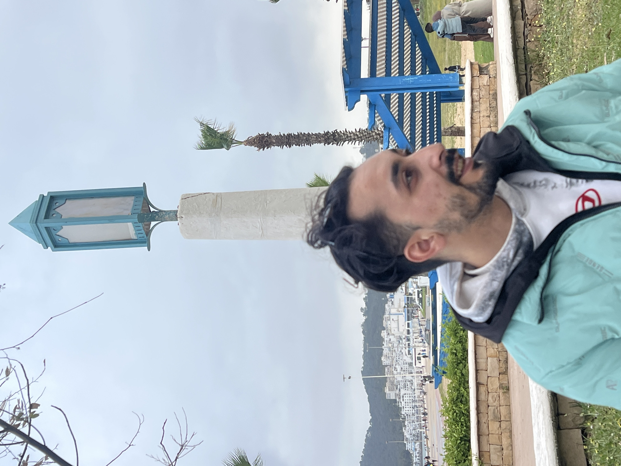 The image is a candid shot taken at eye level, featuring a man in the foreground, situated on a promenade overlooking the city of M'Diq, Morocco.
The man is positioned on the right side of the frame, facing away from the camera. He is wearing a light blue jacket with a black hood over a white t-shirt. He has dark hair and a beard, and his facial expression is one of looking up and smiling.
To the left and behind the man, there is a waterfront view. The city of M'Diq is visible with its white buildings and coastal setting. There is a mountain in the background with trees.
In the midground, elements of the promenade are visible. There is a blue pergola structure on the right side.
The sky is overcast, contributing to the overall light.