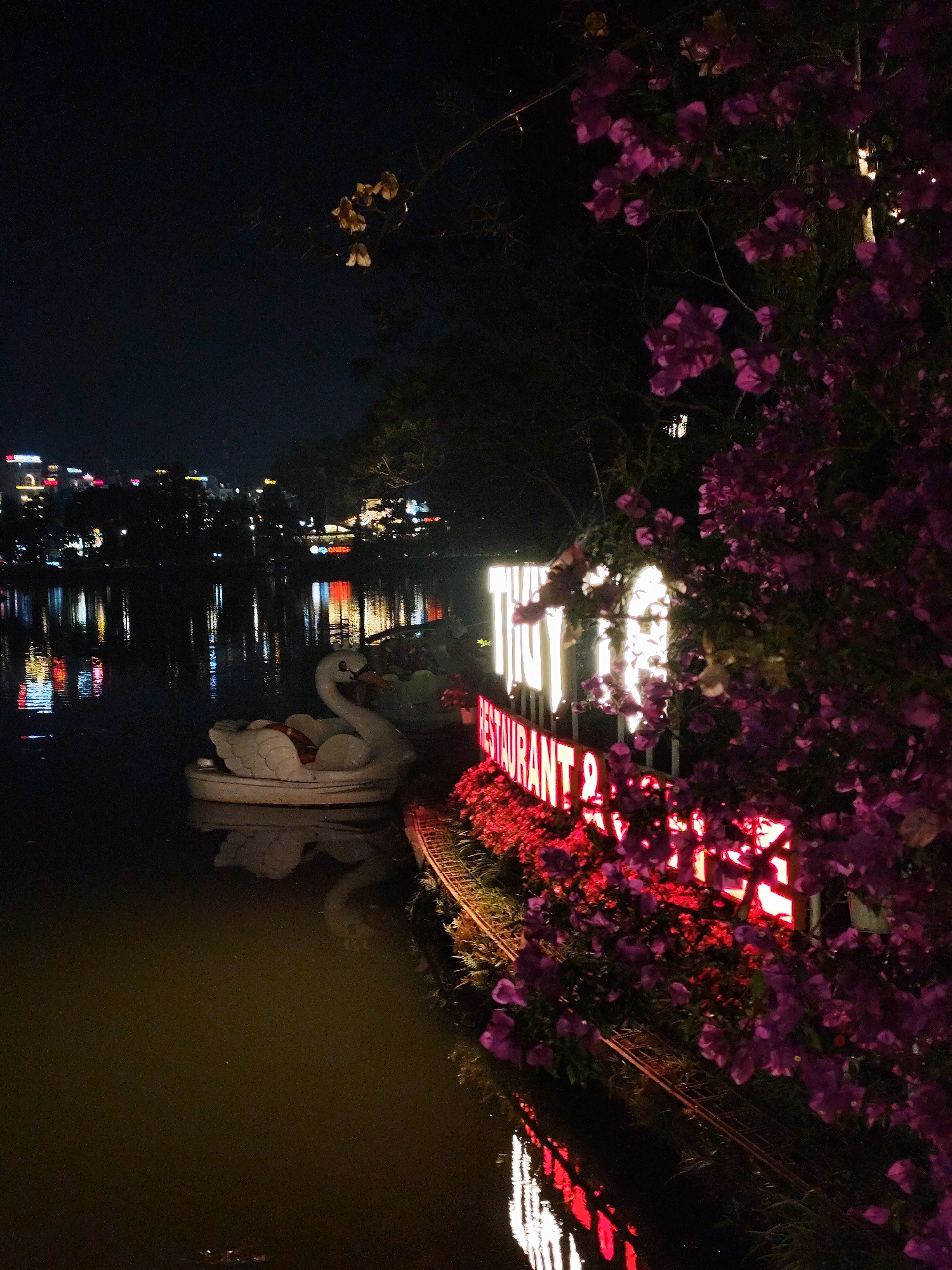 The scene depicts a nighttime view of Xuan Huong Lake in Da Lat, Vietnam. On the right, a dense mass of magenta bougainvillea flowers partially frames two illuminated signs. The upper sign displays "THUY" in large, white glowing letters, while the lower sign reads "RESTAURANT &" in red glowing letters, bordered by a line of red-illuminated foliage along the water's edge. Two white, swan-shaped paddle boats are moored on the dark lake surface near the illuminated shore, reflecting the adjacent lights. Across the lake, the illuminated skyline of Da Lat city is visible in the background, with its diverse lights reflecting on the water. The sky is dark. No people or active interactions are present.