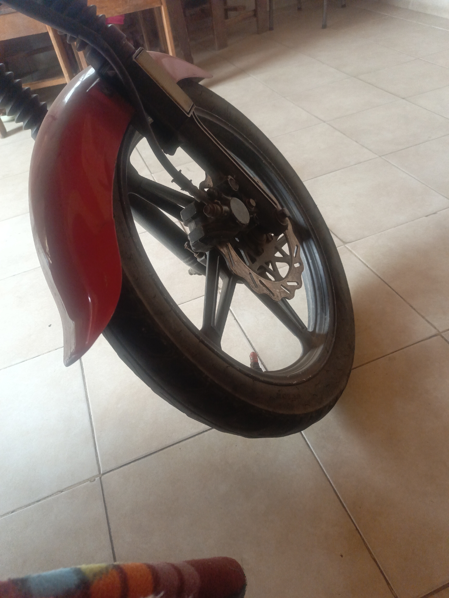 The image shows the front wheel of a motorcycle, specifically the left side, with a red fender. The wheel is made of a black alloy, with a worn tire. A disc brake is visible. The background is a tiled floor, suggesting an indoor location. The presence of chairs indicates a domestic or commercial setting. The location details (Juan Griego, Venezuela) are not directly visible within the image itself.