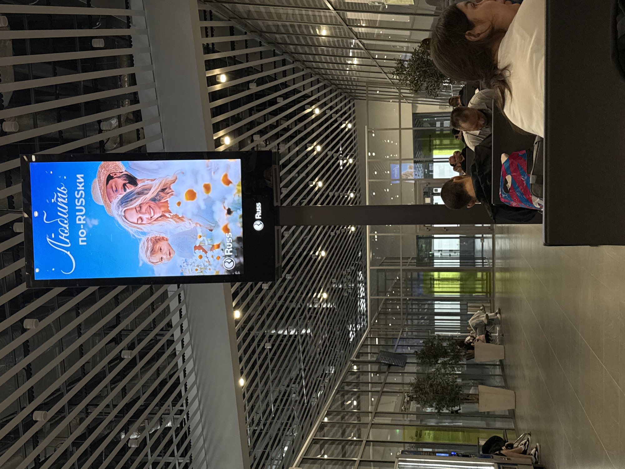 The image captures an indoor scene, likely a modern airport or public transit station, featuring a large digital advertisement screen mounted on a dark gray pillar. The screen displays a vibrant advertisement with Russian text at the top that translates to "Love." Below this text is "po-RUSSki," which can be interpreted as "in Russian style" or "like Russians." The advertisement features a smiling couple and a child surrounded by white and orange flowers. The brand logo "Russ" appears twice on the screen.
The environment is characterized by a high ceiling with a grid of diagonal white beams and numerous circular light fixtures. The floor is tiled in a light gray. Large glass windows line the right side of the image, revealing a glimpse of an exterior or another section of the building, with green and yellow lighting visible in the distance.
Several people are seated in a waiting area in the foreground and midground. In the foreground, a woman with dark hair is looking to the right, wearing a white t-shirt. To her left, a man with short hair is seated, with a backpack featuring a colorful, abstract pattern placed behind him. Further in the midground, other individuals are seated, some appearing to be sleeping or resting. Two large potted plants with dark green foliage stand near the glass windows, adding a touch of greenery to the otherwise modern interior. The overall atmosphere suggests a moment of waiting or transit within a well-lit, contemporary public space. The location being in Boldino, Russia is not ascertainable from the image itself.