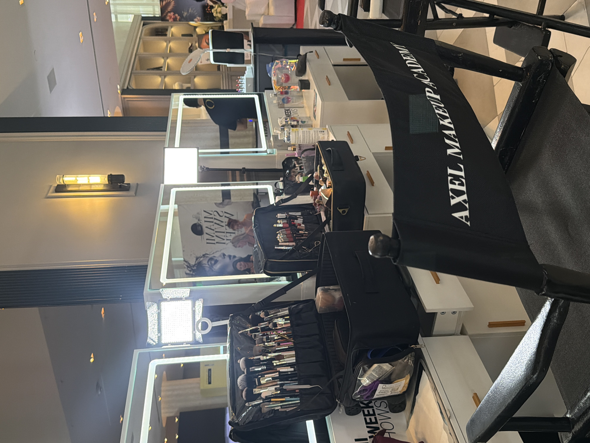 This image captures an interior scene, likely a makeup studio or academy. The foreground is dominated by a black director's chair with "AXEL MAKEUP ACADEMY" printed in white on its back. The chair's canvas seat and frame are also black. In front of the chair, there are several makeup stations, each equipped with a mirror, a bright LED light, and a makeup bag filled with brushes and various cosmetic products. The makeup stations are arranged in a vertical line, receding into the background. In the mirrors, there are reflections of people, possibly makeup artists or students. The reflections also provide a glimpse of the surrounding environment, which seems to be a well-lit, modern interior with white walls and other furniture. The overall setting gives the impression of a professional makeup training environment. It is impossible to ascertain the precise location as being in Miami Beach. However, given the name on the chair and the general impression, a reasonable assumption can be made that the image was taken somewhere in the United States of America.