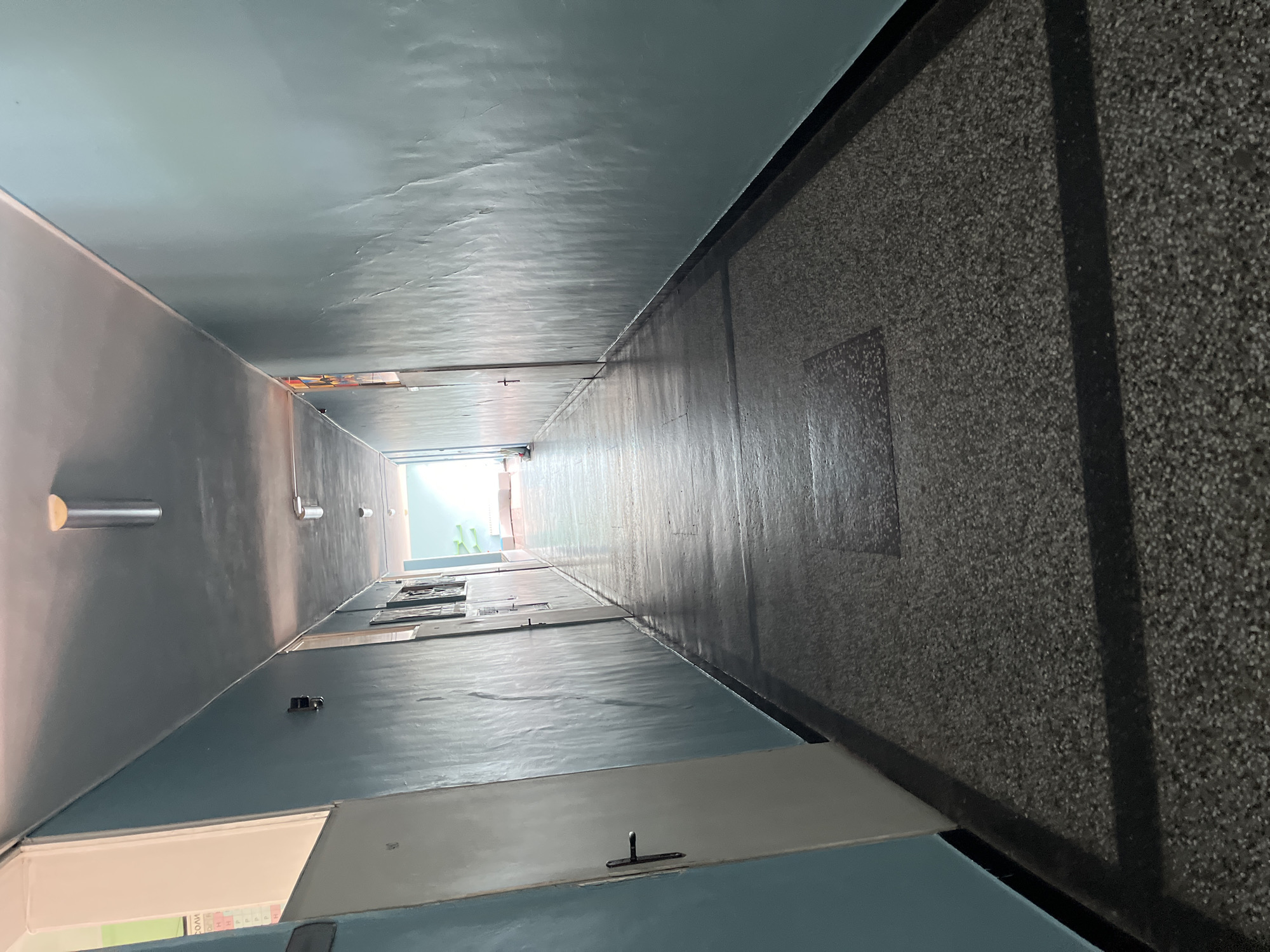 The image shows a deep, narrow stairwell in what appears to be an apartment building, shot from a low angle looking upwards. The walls are painted a pale blue, with doors visible along one side. Lighting fixtures are visible on the left wall. The floor is a speckled gray, contrasting with the smooth, painted walls. Natural light streams in from the top of the stairwell, suggesting an opening to the outside. The location is identified as being in Asenovgrad, Bulgaria, though the image itself does not inherently display identifiers for that city.