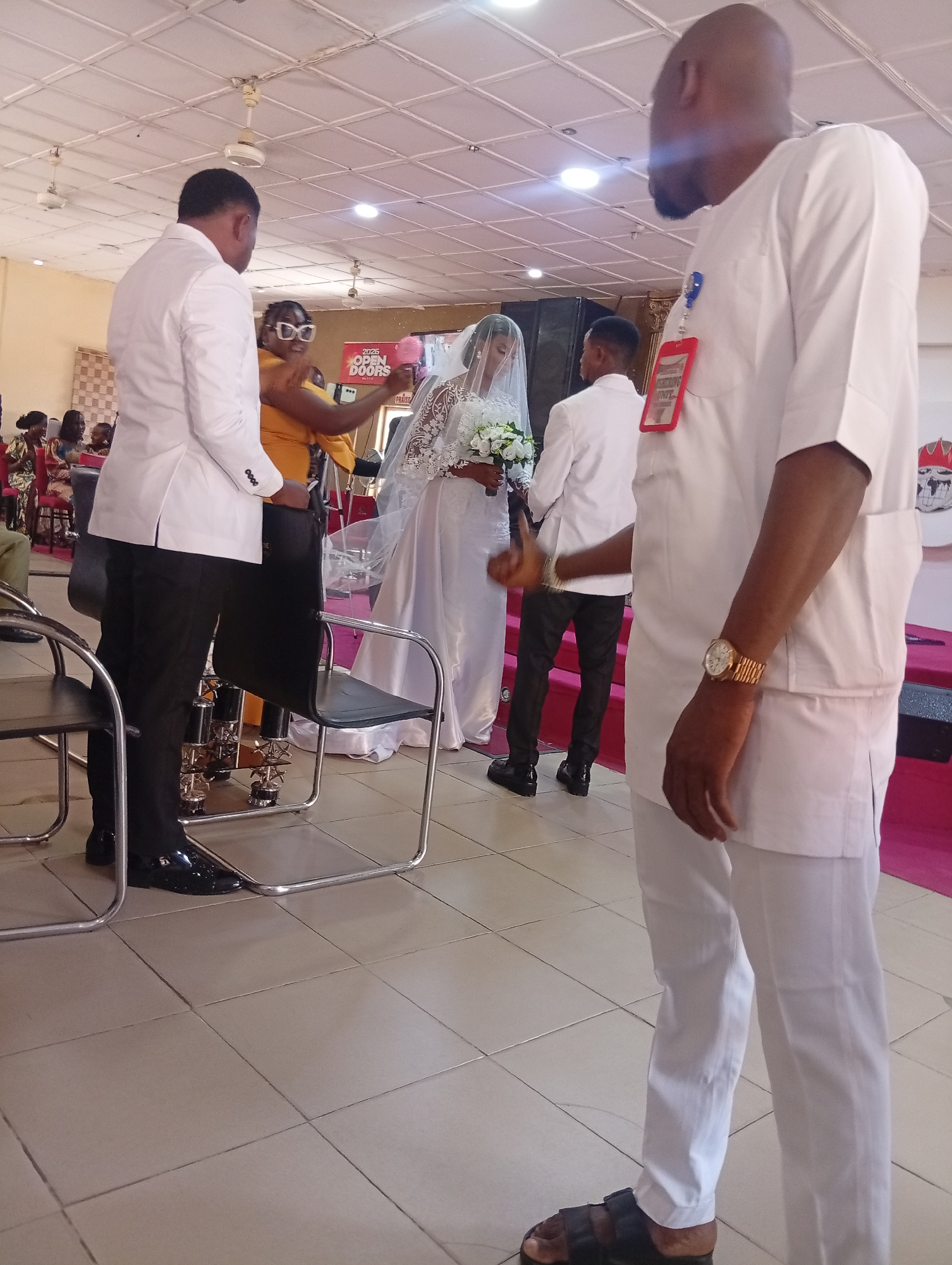 The image depicts a wedding ceremony in Dutse, Nigeria. In the foreground, a man wearing a white traditional attire and black sandals stands facing away from the camera. He is observed to be wearing a gold wristwatch. To his left, a man in a white suit and black shoes is facing the bride and groom. Behind the man in the suit, a woman in an orange dress and glasses is visible, holding a pink object. The bride, wearing a white wedding gown and veil, is holding a bouquet of white flowers and is standing next to the groom, who is dressed in a white suit. In the background, other guests are seated. The event is taking place indoors, with a tiled floor and a suspended ceiling with fluorescent lighting and ceiling fans. A banner with "2025 OPEN DOORS" is visible on the right side of the image.