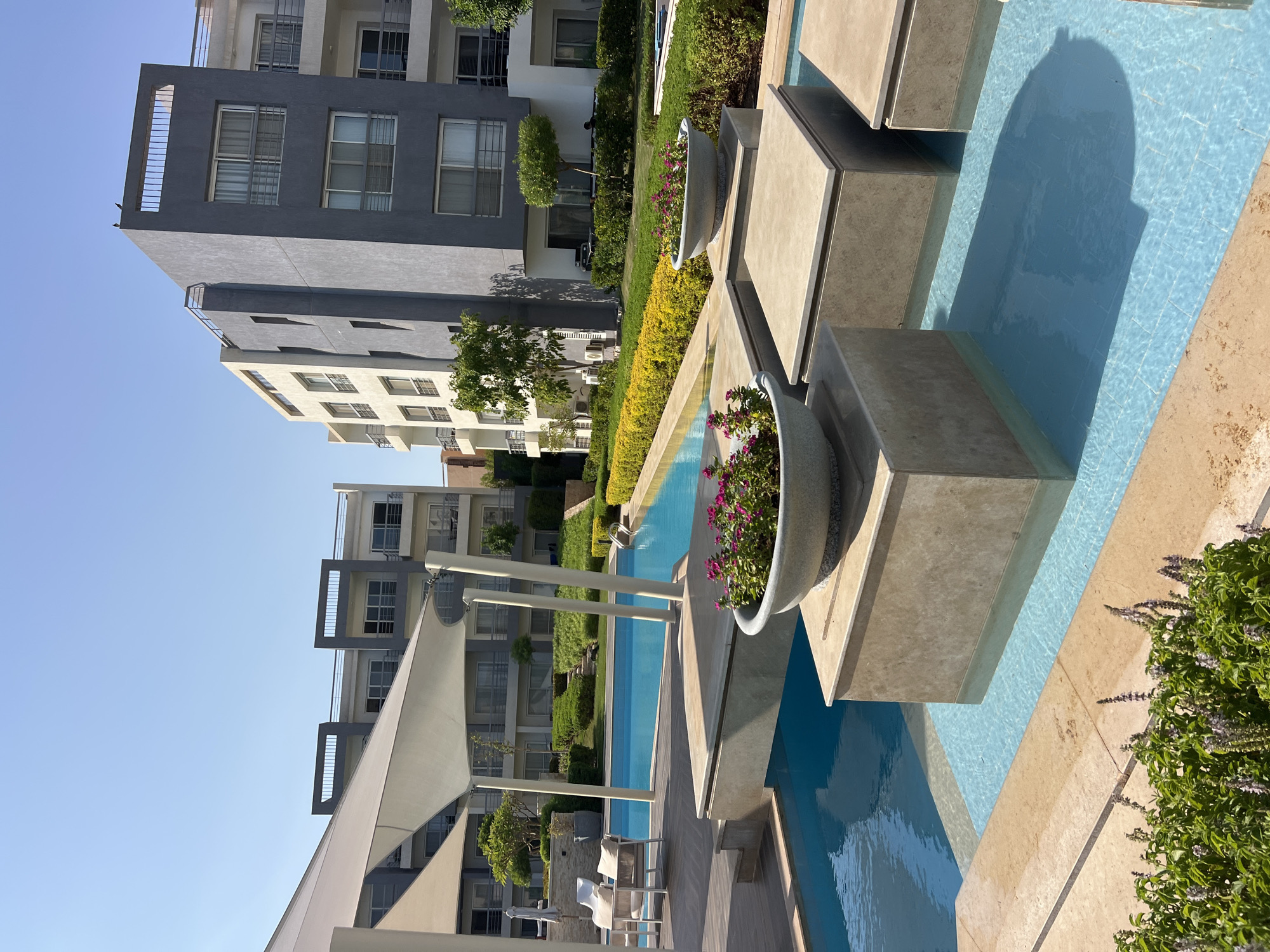 The image depicts an outdoor swimming pool area with modern apartment buildings in the background. The pool is a vibrant turquoise blue and features several submerged rectangular platforms with stone planters containing pink flowers on top. The surrounding area is paved with light-colored stone tiles and features lush green landscaping, including hedges and yellow flowering bushes. A large white fabric shade structure is visible to the left, supported by white poles. The apartment buildings are multi-story structures with a contemporary design, featuring balconies and large windows. The sky is clear and blue. The scene appears to be a resort or residential complex in a sunny, warm climate.