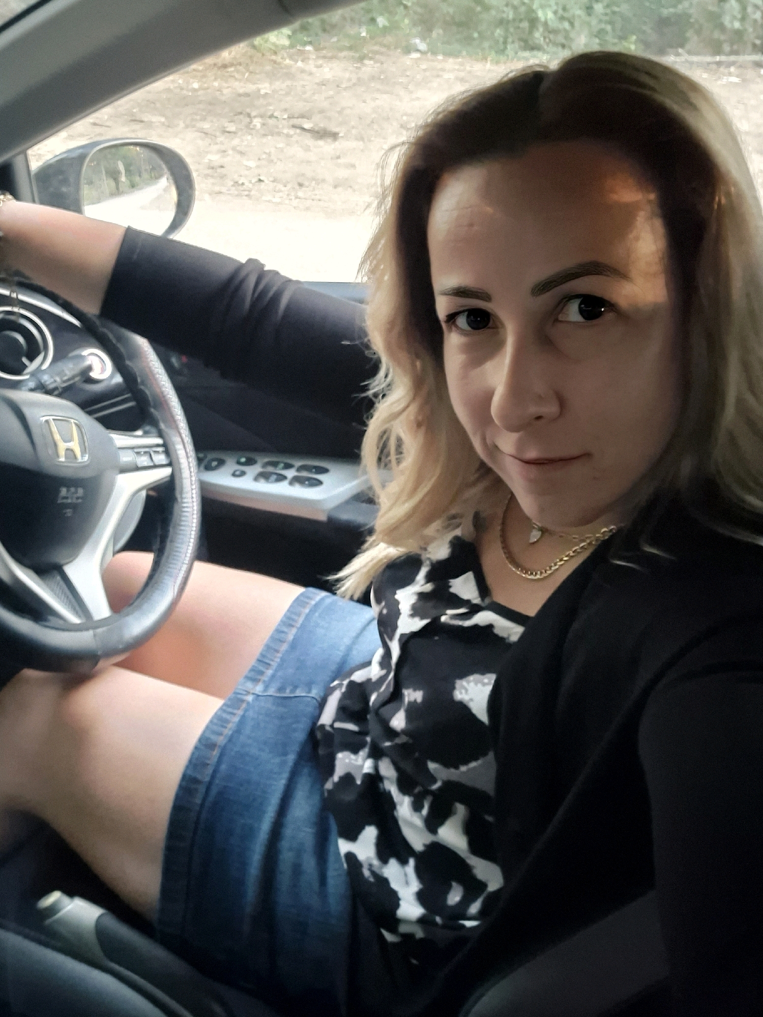 A blonde woman is sitting in the driver's seat of a car. She is wearing a black jacket over a black and white patterned shirt and a denim skirt. The steering wheel is visible in the foreground, with the Honda logo on it. The dashboard and part of the car's interior are also visible. The woman is looking directly at the camera and smiling. The car appears to be parked on a dirt road surrounded by trees.