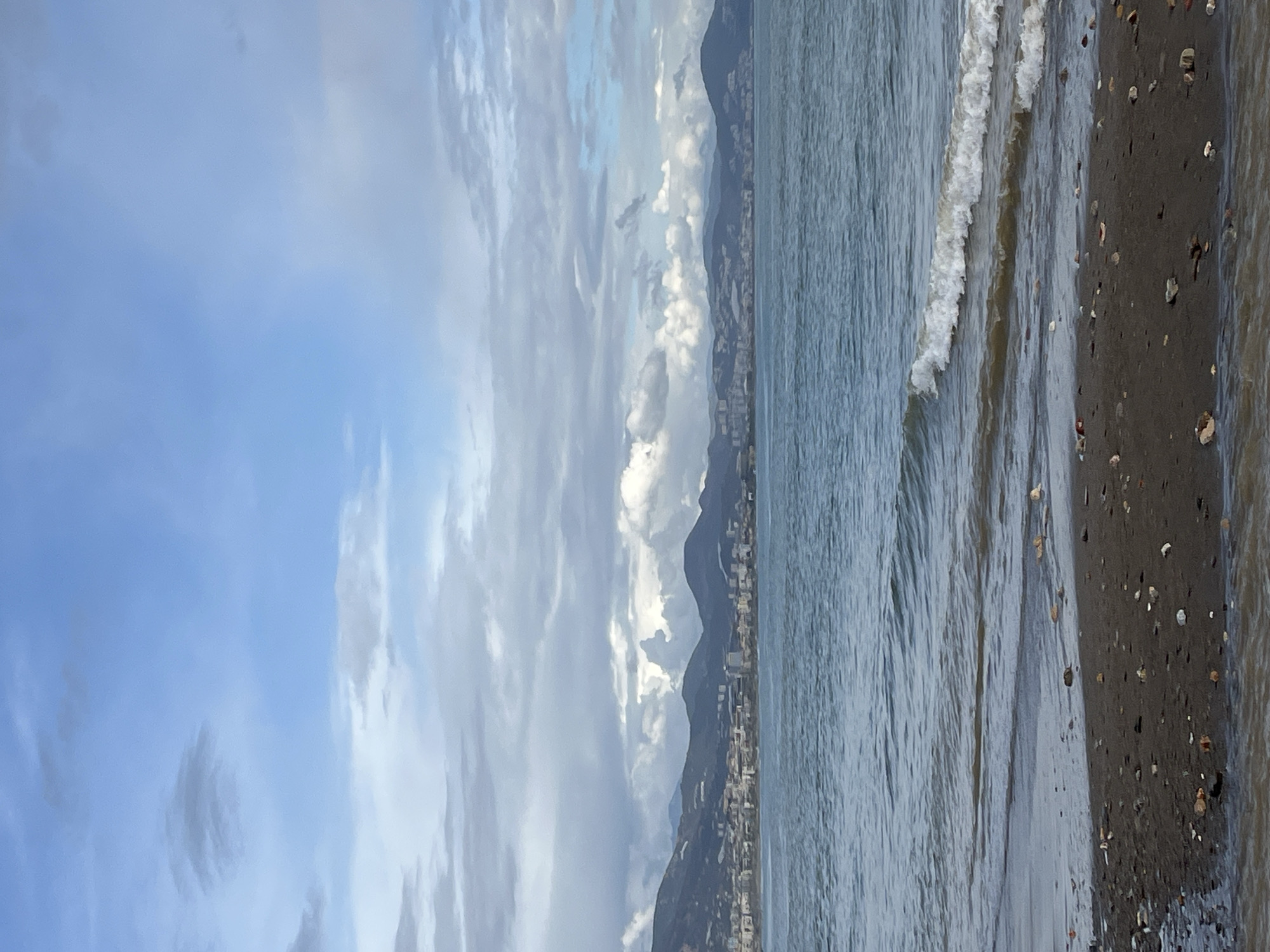 The image presents a seaside view, likely in the city of Alanya, Turkey, given the geography and urban presence along the mountain range in the background. The sky above is covered with scattered, low-lying clouds, varying in hues of white and grey. A mountain range runs along the horizon, with a linear array of buildings visible along its lower slopes, indicating urban development. The ocean occupies the middle ground, with gentle waves approaching the shore. The shoreline is dark, sandy, and interspersed with pebbles and small rocks. There are no people visible in the shot.