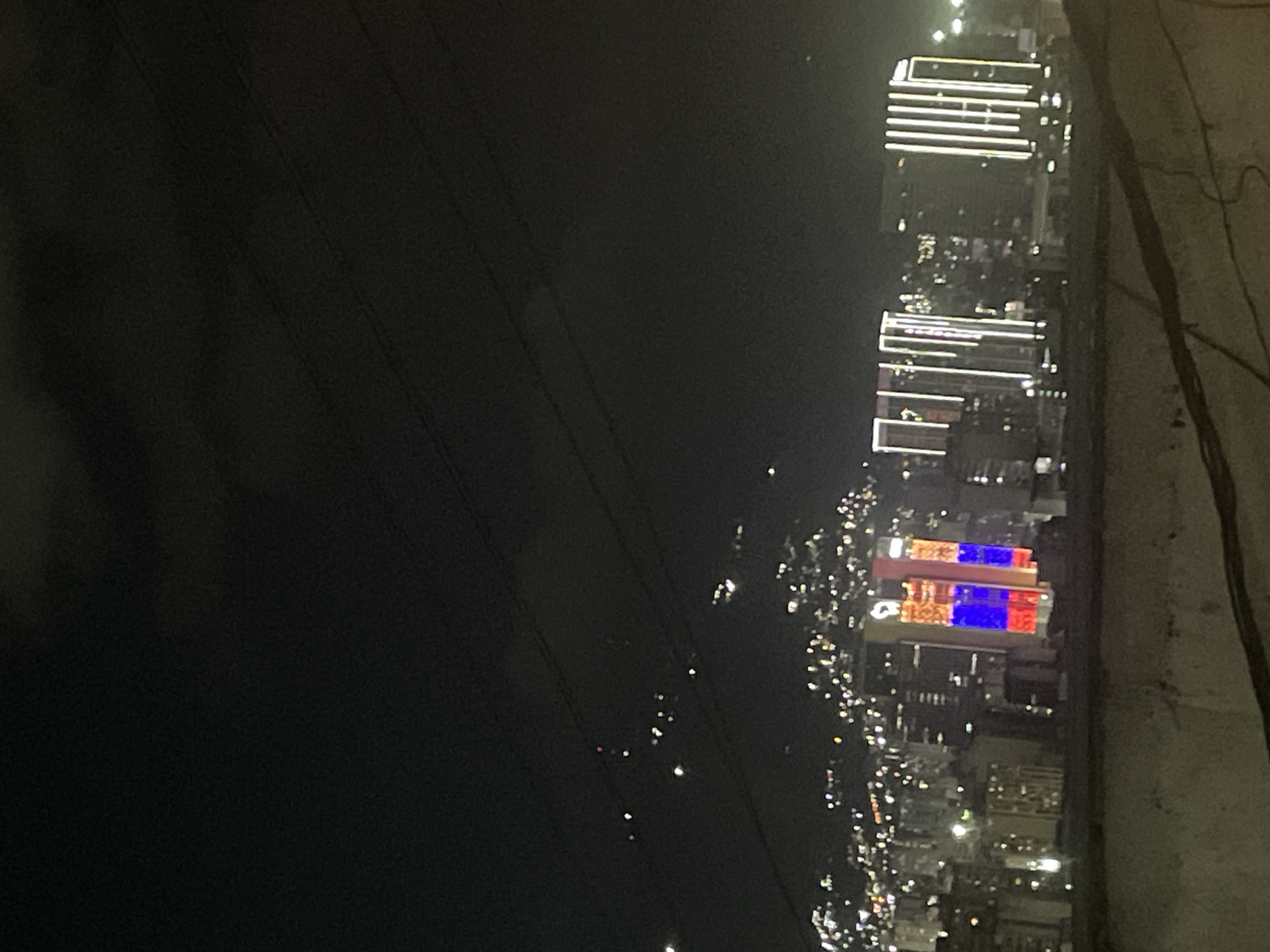 The image depicts a nighttime view of the city of Caracas, Venezuela. Buildings are illuminated with white and colored lights, with some buildings showing a pattern of red and blue lighting. The scene is viewed from an elevated position, potentially a building or hillside overlooking the city. Overhead power lines stretch across a portion of the left side of the image. The right side shows a wall of a building or a bridge with some dangling wires. The background on the left is very dark.