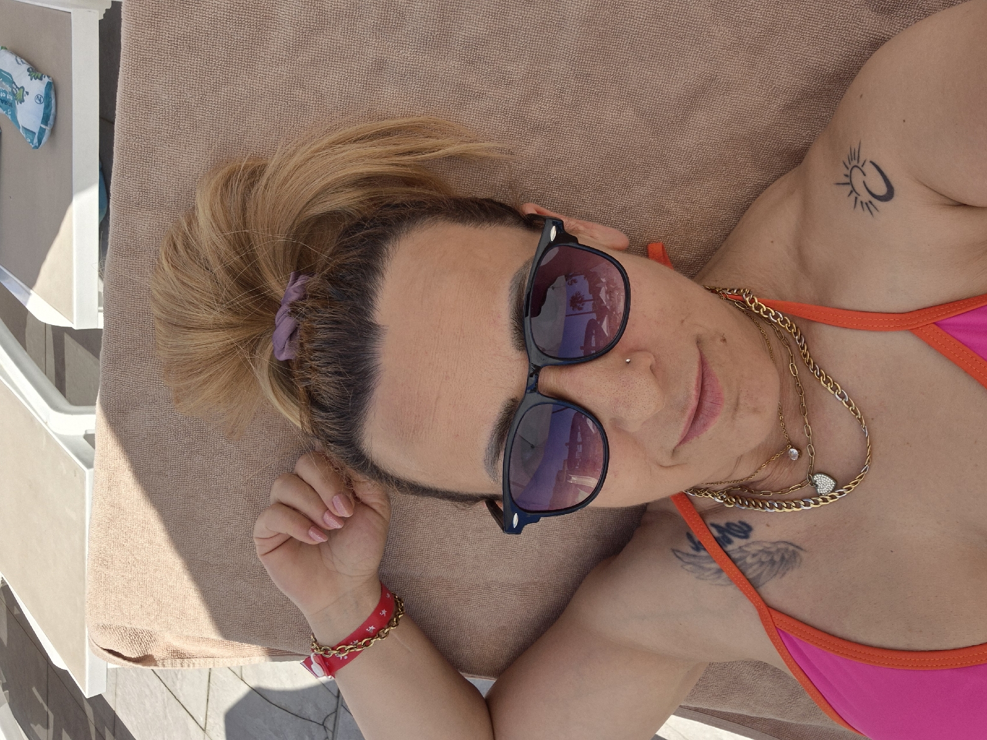 A woman is lying on a lounge chair on a towel outdoors. She is wearing sunglasses and a pink bikini top with orange trim. Her hair is pulled up in a high ponytail. She has several necklaces on, and a red and gold bracelet on her left wrist. She has tattoos on her left shoulder and her right shoulder. The background shows lounge chairs and paving stones.