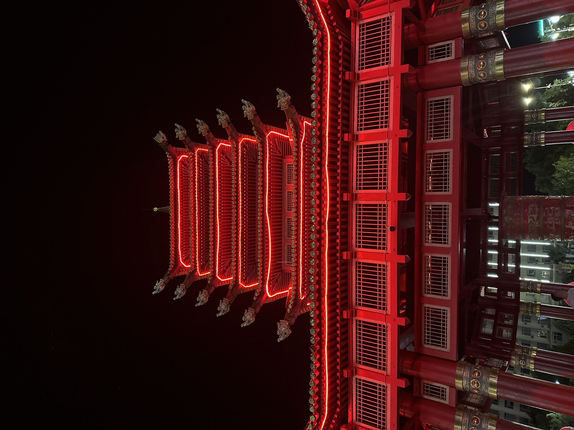 The image captures a brightly lit, multi-tiered pagoda-style building at night. The structure is predominantly red, adorned with glowing red neon lights outlining its multiple roofs and decorative elements. The pagoda has a distinct traditional East Asian architectural style, featuring upturned eaves, each decorated with what appears to be a dragon head ornament. Below the pagoda, a red, lattice-like structure is visible, supported by thick red pillars decorated with gold accents. In the background, indistinct buildings and streetlights suggest an urban environment. The overall scene is vibrant and festive, likely a cultural landmark. The location identified is in the city of Elista, Russia. While the image shows no specific actions, the presence of a few people in the lower portion of the frame, partially obscured, indicates a public space.