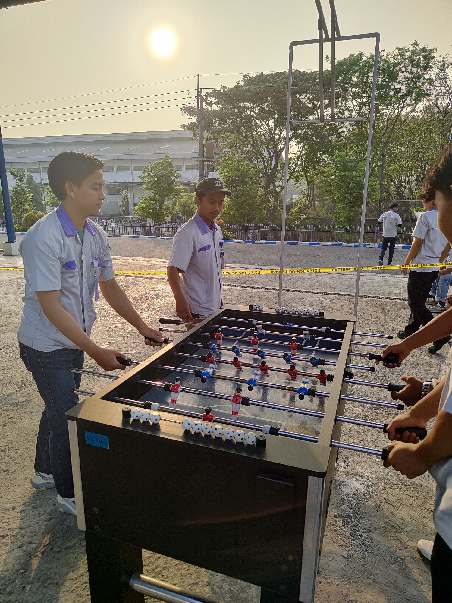 Two young men are playing foosball outdoors. The man on the left, wearing a light gray polo shirt with purple trim and blue jeans, is actively playing. The man on the right, wearing a similar shirt and a black baseball cap, is also playing. They are positioned at opposite ends of a black foosball table. Several other individuals are visible in the background, some standing and observing. The scene appears to be an industrial or factory area, with a large building and trees in the background. A yellow caution tape is strung across a fence in the midground, suggesting a restricted area. The lighting indicates it is late afternoon or early evening, with the sun visible in the hazy sky. The overall environment is an outdoor paved area with some gravel. The location is identified as Raci, Indonesia.