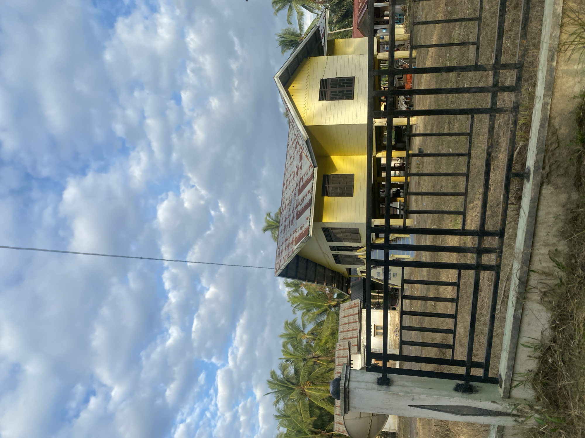 The image depicts a two-story yellow house with a metal roof, partially obscured by a black metal fence. Coconut trees are visible behind the house. The sky is cloudy with patches of blue. The location appears to be in Kruengbeukah, Indonesia. There are no people visible in the image. The environment seems to be a residential area.
