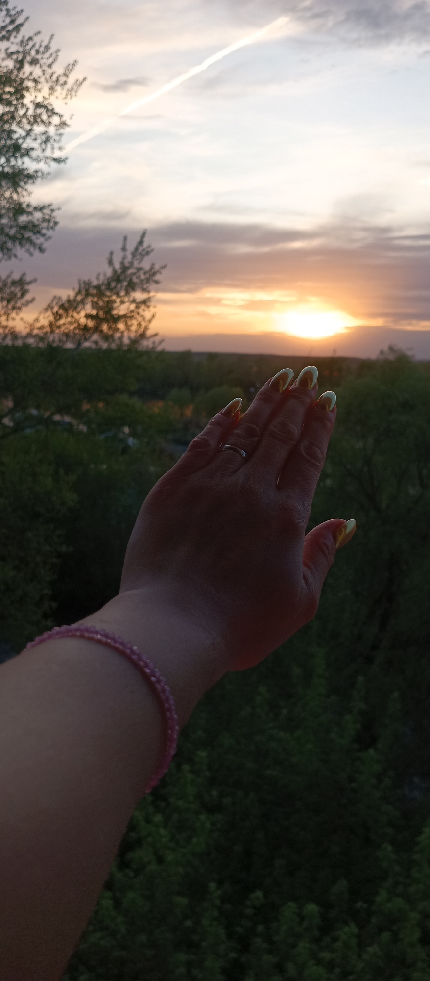 A person's hand is positioned in the foreground, appearing to reach toward the sunset. The hand has long, light-colored nails, and a ring is visible on one finger. A pink beaded bracelet is on the wrist. The background consists of trees and a sunset with a bright sun partially obscured by the horizon. A contrail is visible in the sky. The location is unclear; the city is unknown.