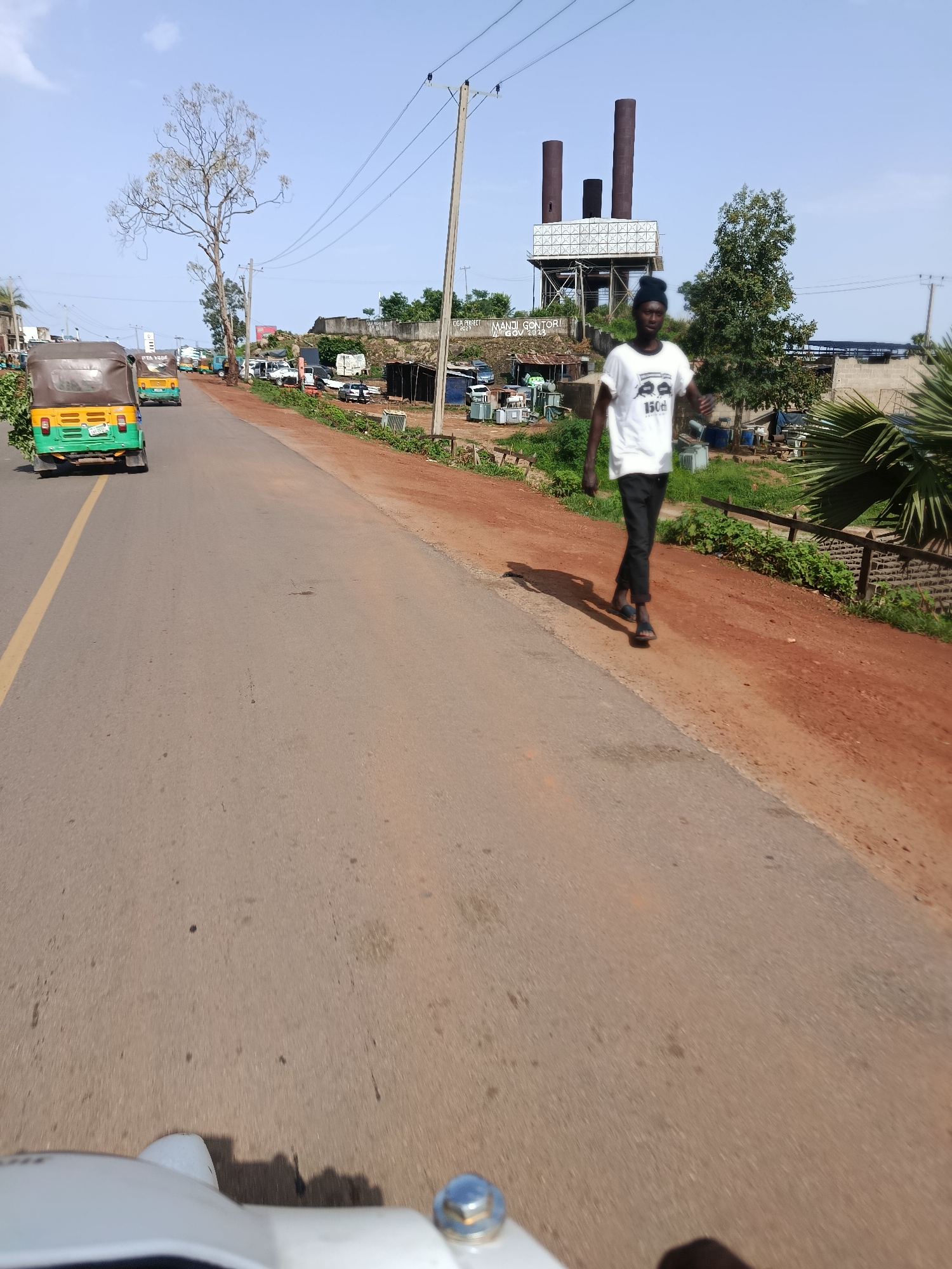 The image depicts a street scene in Rafin Par, Nigeria, under a clear blue sky. In the foreground, the front of a white vehicle is visible. The road, paved with asphalt, has a yellow line marking the lane. On the left side of the road, a green and yellow auto-rickshaw, also known as a 'keke napep,' is driving away from the camera. Further down the road, another similar vehicle is visible.
To the right of the road, a man with dark skin is walking. He is wearing a white t-shirt with a graphic design and dark pants, along with black sandals. He is looking towards the right side of the frame. The ground beside the road is a reddish-brown dirt. In the background, there are various structures, including a tall industrial-looking tower with three smokestacks, a water tower, and some low-rise buildings and sheds. Vegetation, including trees and bushes, is scattered throughout the scene. Power lines run across the sky, supported by utility poles. The overall impression is that of a busy, somewhat industrial area on the outskirts of a city.