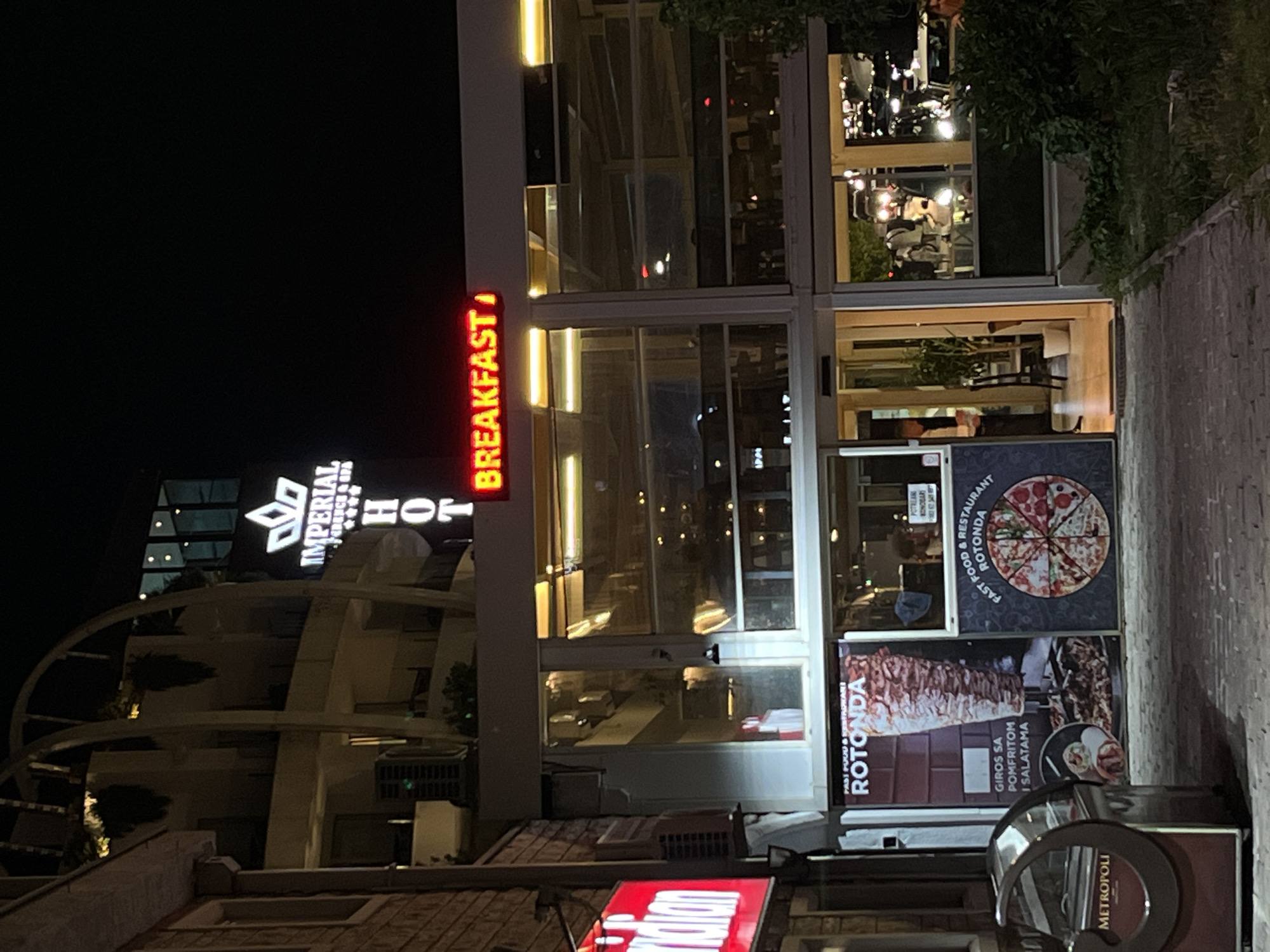 The image shows the exterior of a building at night, likely in Budva, Montenegro, identified by the "IMPERIAL RESIDENCE & SPA" sign. The building features a restaurant with a bright red sign that reads "BREAKFAST" in capital letters. Below the "BREAKFAST" sign, there are windows offering a view into the restaurant, where tables and chairs are visible. To the left of the restaurant entrance, there is a red sign with white lettering that appears to say "indian". Further left, a circular display case with "METROPOLI" written on it is present. The entrance to the restaurant is a glass door, and to the right of the entrance, there are posters advertising "ROTONDA FAST FOOD & RESTAURANT" with images of a döner kebab and a pizza. A tree and some green foliage are visible to the right of the building, and streetlights or building lights illuminate the scene. The pavement in front of the restaurant is made of cobblestones.