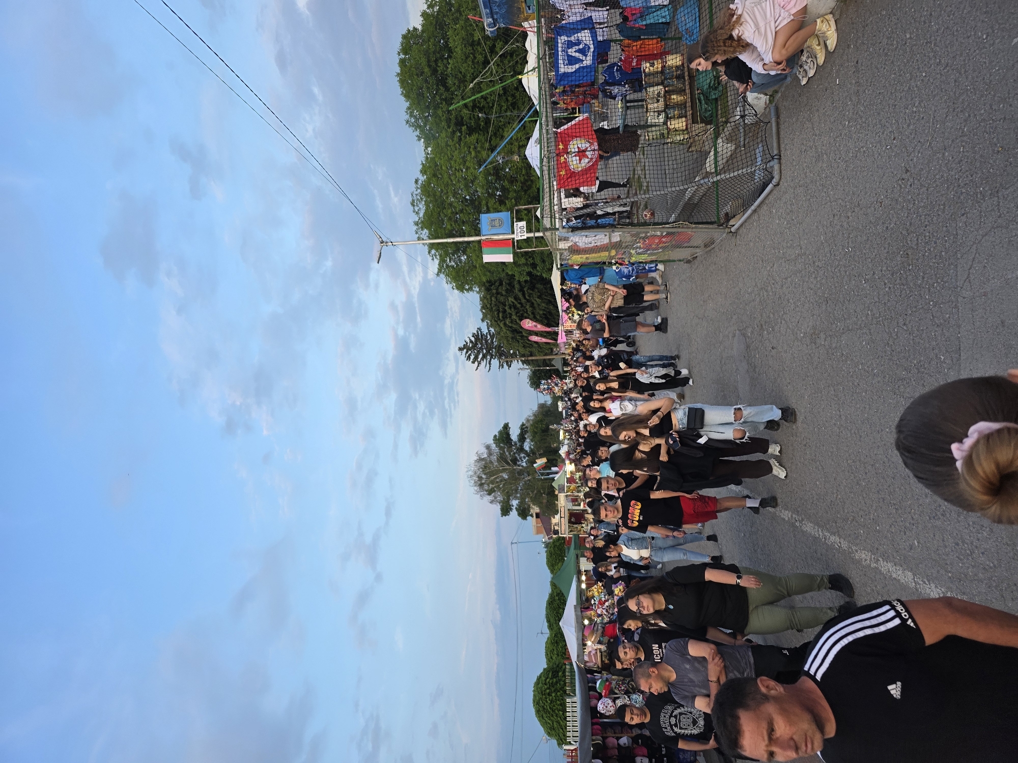 The image depicts a crowded outdoor market or festival taking place on a paved street in Elin Pelin, Bulgaria, during the late afternoon or early evening. A multitude of people, primarily young adults and some middle-aged individuals, are walking along the street, browsing stalls, or standing.
On the left side of the street, multiple vendor stalls with white canopies are visible, displaying merchandise such as hats and clothing. On the right side, stalls are constructed using metal mesh fences, showcasing various items including hanging t-shirts, jerseys, and two distinct flags: a red flag with a white star and emblem, and a blue flag with a white "WA" logo. Further down this side, shelves hold an assortment of jars or containers. Two young women are observed sitting on the curb next to one of these right-side stalls.
In the mid-ground, a prominent street light pole displays two flags: the national flag of Bulgaria (white, green, red horizontal stripes) and a blue flag with an emblem, likely representing the municipality of Elin Pelin. Below these flags, a white sign with the number "100" is affixed. Overhead, dark electrical wires traverse the sky.
The street surface is asphalt, and mature trees line the background. The sky is predominantly blue with scattered, light grey clouds, indicative of a clear but slightly overcast atmosphere. The overall impression is one of a bustling community event or fair.