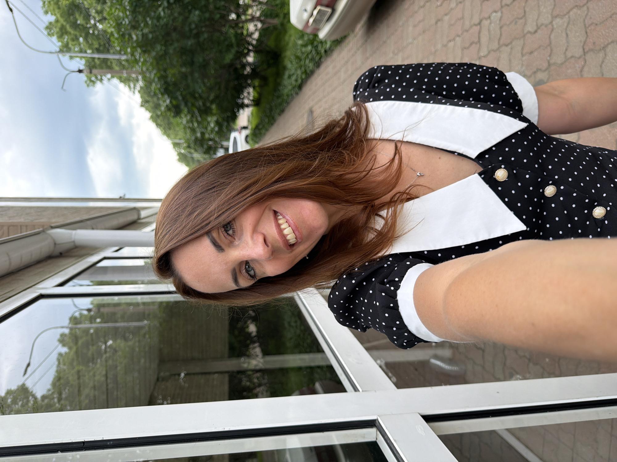 A medium-shot, selfie-style photo shows a woman with shoulder-length auburn hair, wearing a black dress with white polka dots and a large white collar. She is standing outside a building with a reflective glass facade, possibly in Birobidzhan, Russia, as suggested by the metadata. The woman is smiling at the camera, her right arm extended to take the photo. To her right, a paved walkway runs alongside a grassy area with trees and a parked car. The sky is overcast.