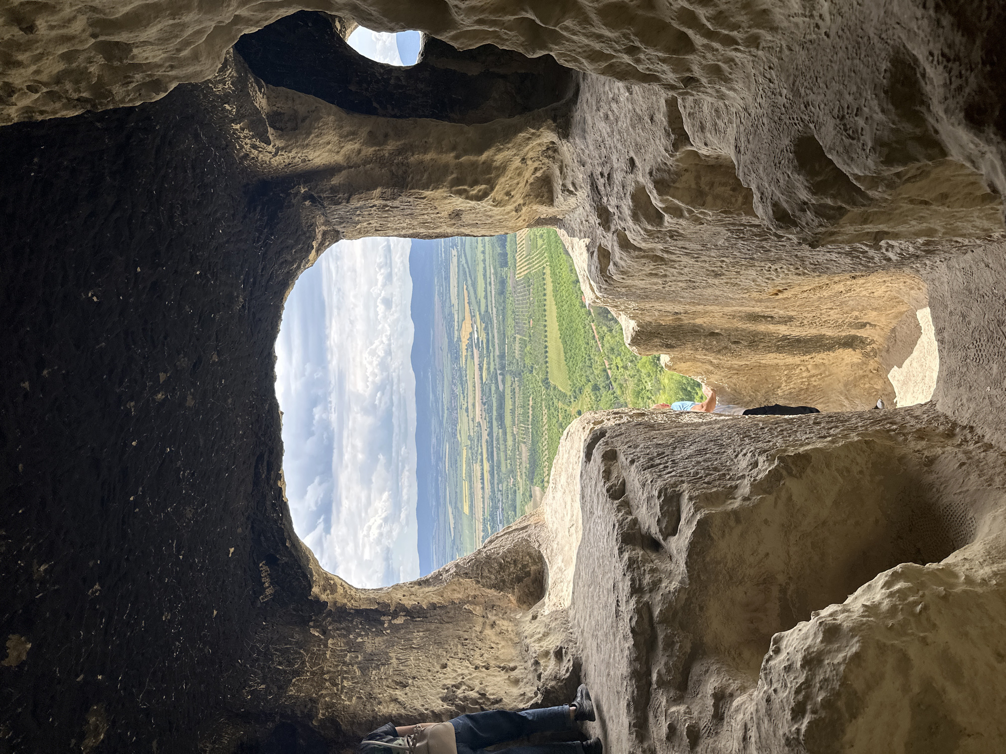 The image shows the view from inside a cave or rock shelter, looking out over a landscape. The cave entrance is roughly oval-shaped, framing a vista of green fields, rolling hills, and a sky with scattered clouds. The interior of the cave is rough, uneven rock, with variations in color from dark to light brown. A person is visible just outside the cave in the upper right of the frame, lying down on the rock. Lower in the frame, another person's legs can be seen. The location is near the village of Milanovo, Bulgaria.