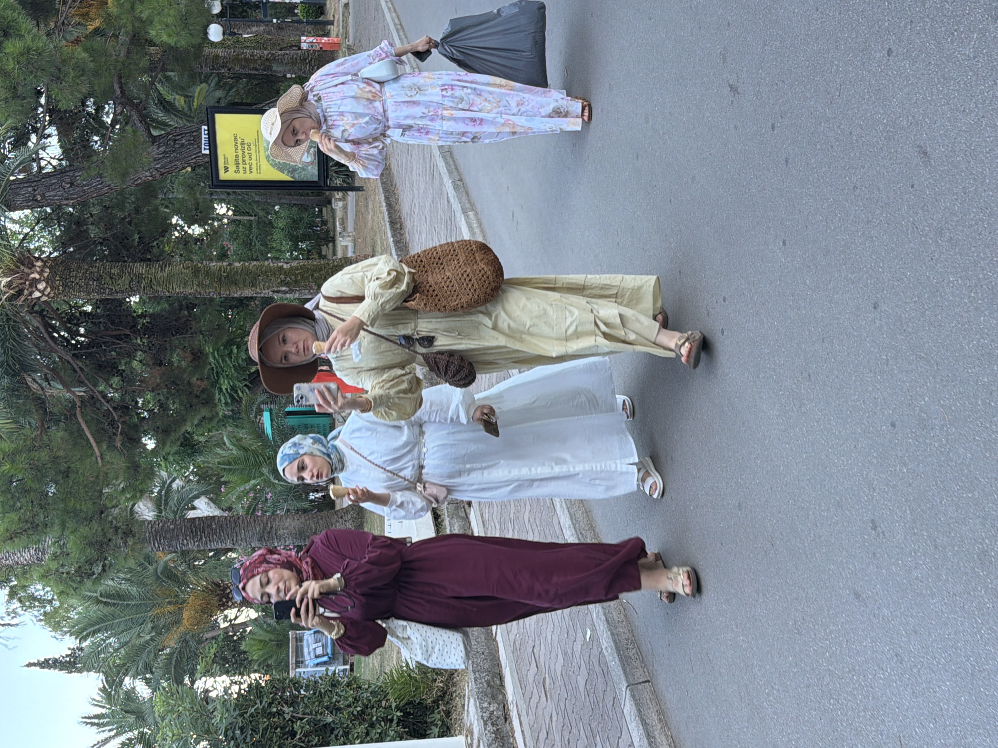 In Budva, Montenegro, four women walk along a paved road lined with palm trees and other lush greenery. The woman on the far left wears a dark maroon abaya and a matching hijab, holding a smartphone in her right hand. Next to her, a woman in a white abaya and a light blue and white patterned hijab holds an ice cream cone in her left hand. The third woman from the left is dressed in a light yellow abaya and a brown hat, also holding an ice cream cone. She has a large woven bag slung over her shoulder. The woman on the far right wears a light-colored floral print abaya and a wide-brimmed straw hat, holding an ice cream cone to her mouth with her right hand and a large black garbage bag in her left. A yellow sign with white text, presumably an advertisement or public notice, is visible in the background behind the women.