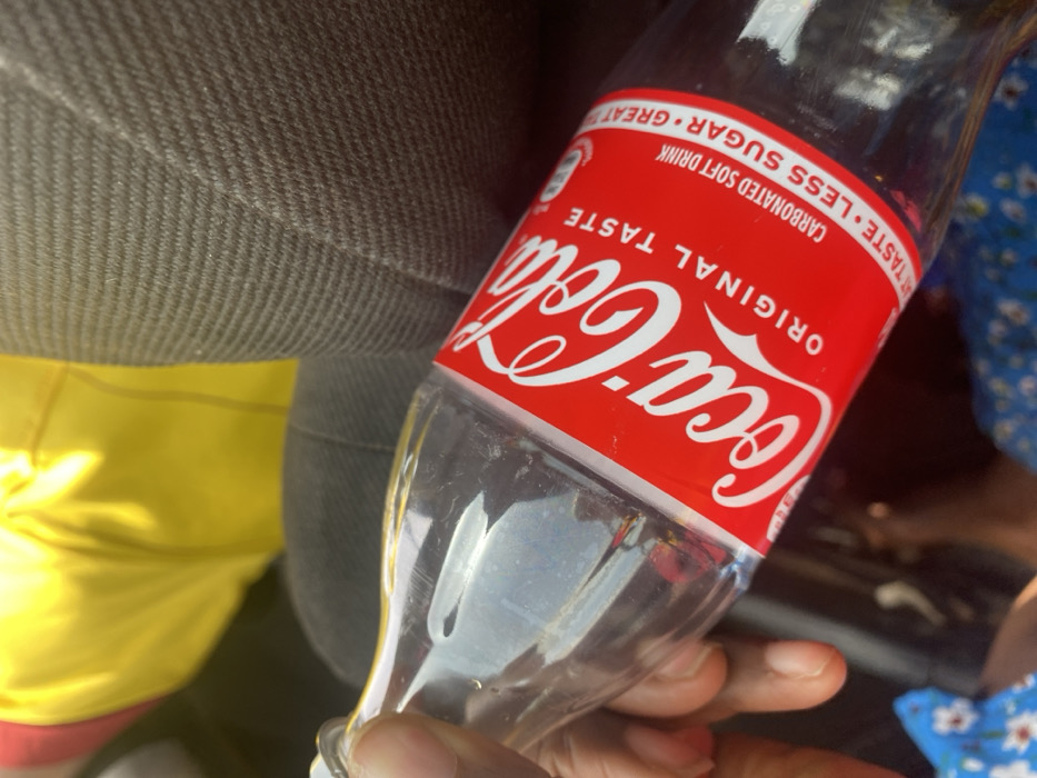 A human hand with dark skin tone holds a mostly empty, clear glass Coca-Cola bottle diagonally across the frame. The bottle features its standard red label, displaying "Coca-Cola ORIGINAL TASTE" and text indicating "CARBONATED SOFT DRINK" and "LESS SUGAR". The background consists of dark grey, ribbed fabric, characteristic of vehicle upholstery, occupying the upper right quadrant. A vibrant yellow fabric is partially visible in the upper left, while a blue fabric with a white floral pattern is visible in the lower left and bottom. The scene is situated in Unguwar Gaya, Nigeria.