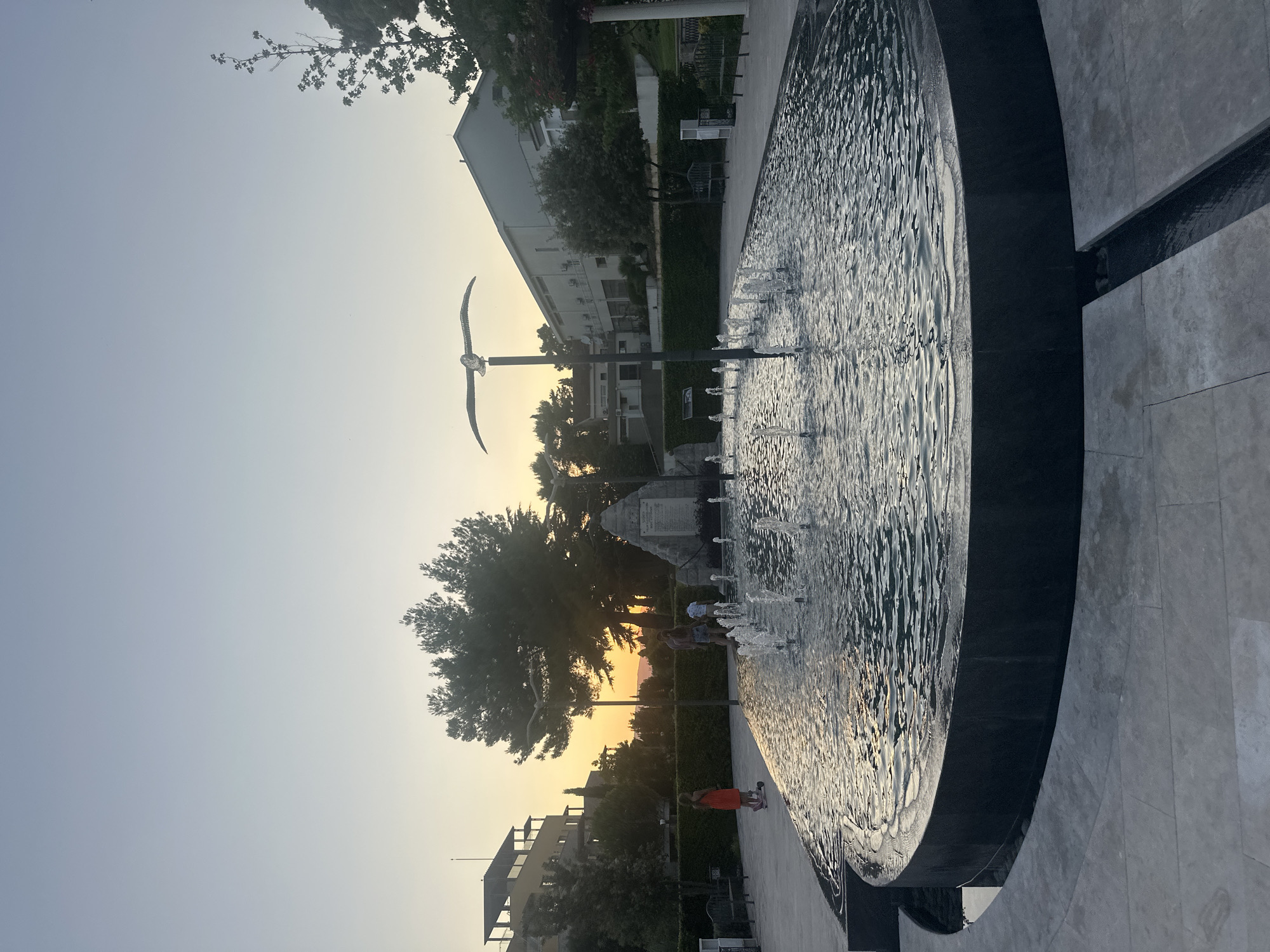 In Kumbor, Montenegro, at dusk, a large, oval-shaped public fountain with multiple jets sprays water into the air. The fountain's edge is constructed of dark, polished stone, and it is surrounded by light-colored paving stones. In the background, a monument with inscriptions stands behind the fountain. To the left of the monument, a cluster of trees is visible, and beyond them, buildings with layered architecture and balconies are situated. On the right side of the fountain, a long, light-colored building with several windows and a sloping roof is present, bordered by a manicured hedge. The sky displays a gradient of pale blue to warm yellow, indicating the setting sun. A metal sculpture of a bird in flight is mounted on a pole near the fountain. A young girl on a scooter is positioned on the left side of the paved area, and two adults and a child are walking near the monument.