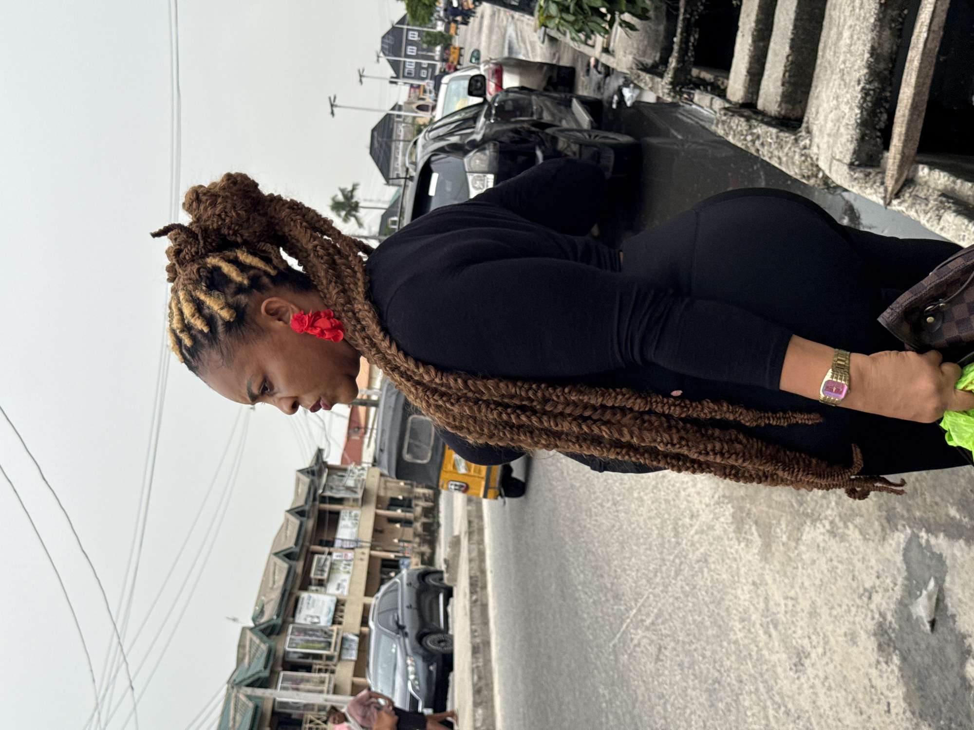 A person is standing on a sidewalk beside a busy street in Ado, Nigeria. The person is wearing a black outfit and has braided hair adorned with red flowers. They are wearing a gold watch on their left wrist and carrying a brown checkered purse. The sidewalk is concrete and appears slightly dirty. The street is filled with cars, including a black SUV and a yellow tricycle. Buildings with green roofs and various signs are visible in the background. Overhead power lines are also present.