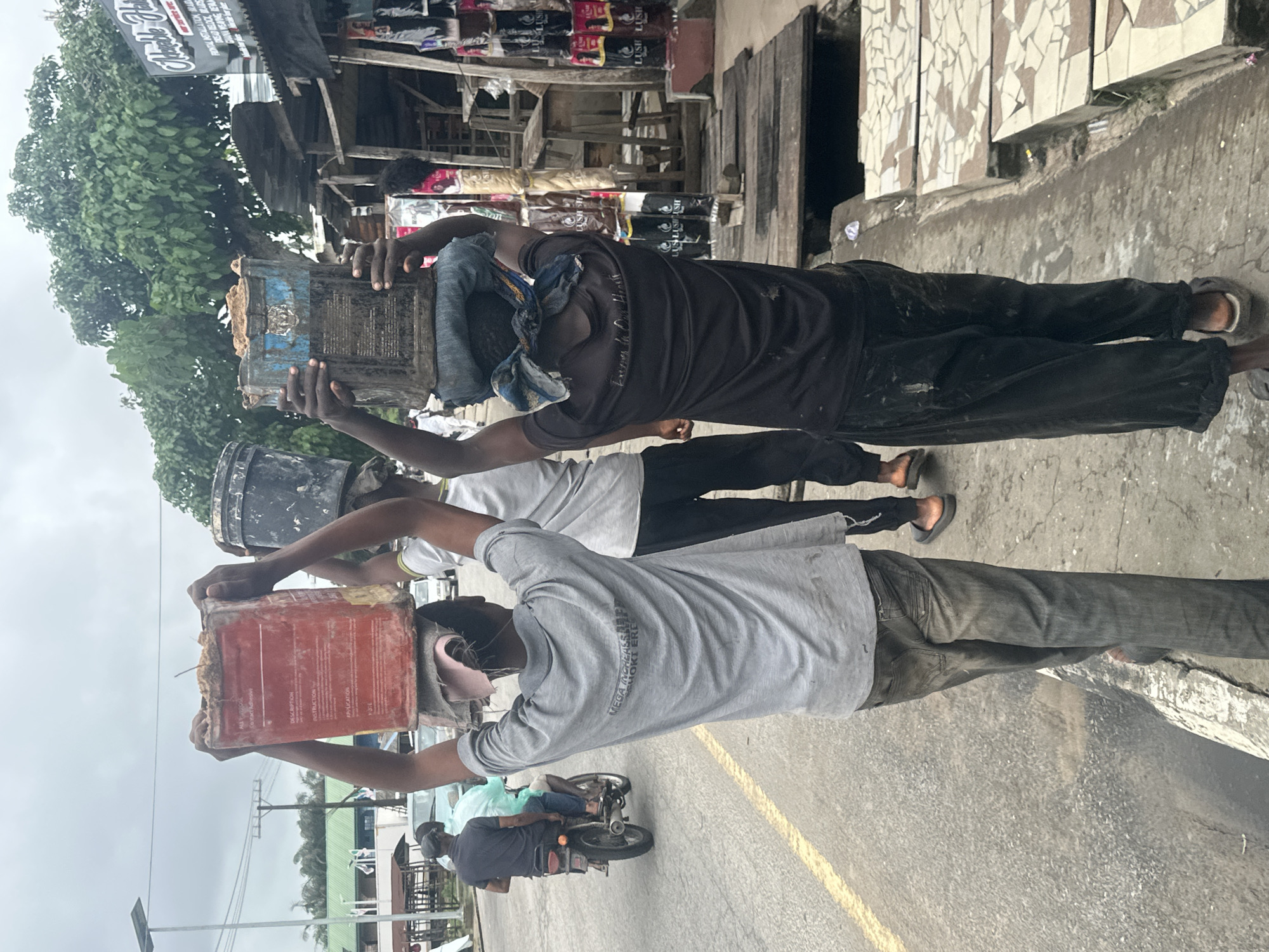 The image shows three people carrying large containers on their heads in what appears to be a street market. The person in the foreground is wearing a grey t-shirt and dark pants, carrying a red rectangular container. To their left, another person in a black t-shirt and black pants is carrying a blue and black bucket. A third person, partially visible in the background, is carrying a dark bucket.
The setting seems to be an outdoor market, possibly in Nigeria, given the "Badagry, Nigeria" location context. There are stalls and structures in the background, with some goods displayed, including what appears to be hair products with "LUSH LUSH" text visible. The ground is paved, and there's a yellow line on the road, suggesting it's a street. The sky is overcast, indicating cloudy weather. The overall mood seems to be one of work and labor, as the individuals are engaged in carrying heavy loads. The text visible includes "DESCRIPTION," "PESTRUCTI," "APPLICATION," "HOKI ERE," and "MEGA INGHEASSLEE."