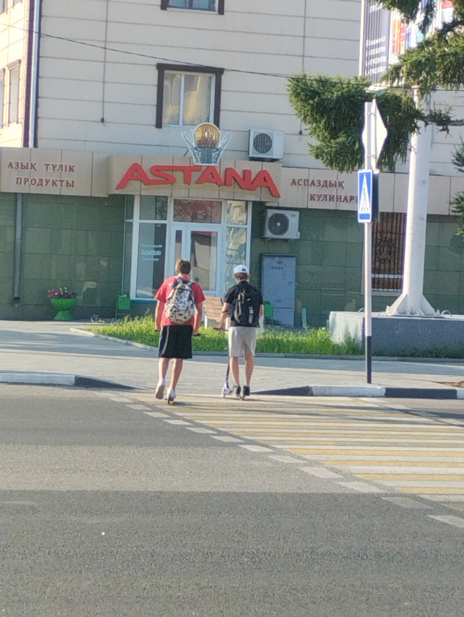 The image shows a street scene in Kostanay, Kazakhstan, featuring a building with signage in Kazakh and Russian. On the left side of the building, the sign reads "АЗЫҚ ТҮЛІК ПРОДУКТЫ" which translates to "Food Products." To the right of this, a large red sign prominently displays "ASTANA," the former name of the capital city, which was renamed Nur-Sultan in 2019 and then reverted to Astana in 2022. Further to the right, another sign reads "АСПАЗДЫҚ КУЛИНАРИ" which translates to "Culinary."
In the foreground, two individuals are crossing a pedestrian walkway. The person on the left is walking and wearing a red t-shirt, black shorts, and a backpack with a patterned design. The person on the right is riding a scooter, wearing a dark t-shirt, light-colored shorts, and a black backpack. Both individuals are seen from behind, facing away from the camera. The environment includes a paved road with a pedestrian crossing, a sidewalk, and a building with green lower walls and light-colored upper walls. Several air conditioning units are visible on the building's facade. A traffic sign indicating a pedestrian crossing is also present. A large green planter with flowers is situated to the left of the building.