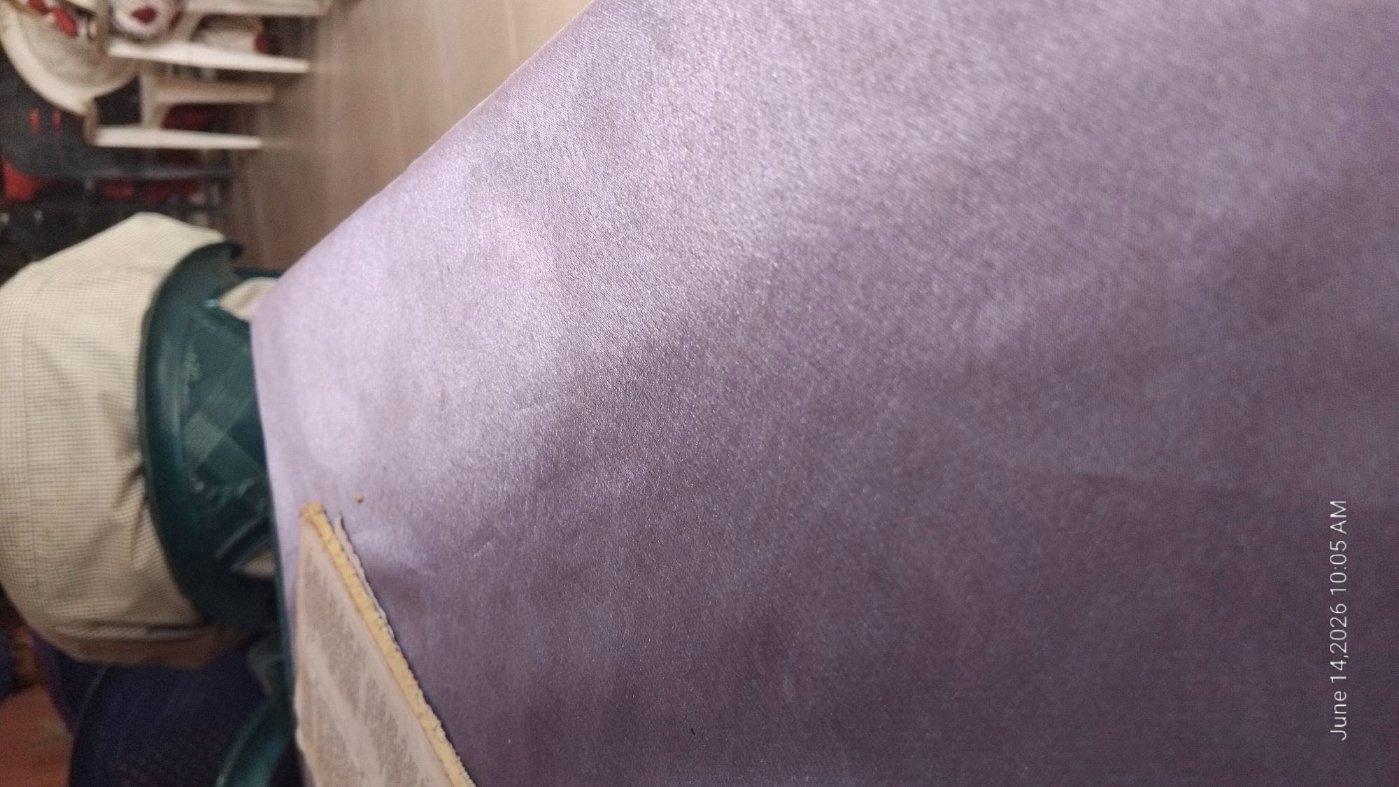The image depicts a close-up view of a purple fabric surface, possibly a tablecloth or a draped cloth, with a slightly reflective sheen. In the upper left corner, a portion of a document with text is visible, lying on the purple surface. In the background, out of focus, are several people seated on chairs. One person is wearing a light-colored checkered shirt. The chairs appear to be dark green and white. The environment suggests an indoor setting, possibly a meeting room or a dining area, with tiled flooring visible in the background. The timestamp "June 14, 2026 10:05 AM" is overlaid on the bottom of the image. The location of Jalingo, Nigeria, is not visually represented in the image itself, and therefore cannot be determined from the visual content.