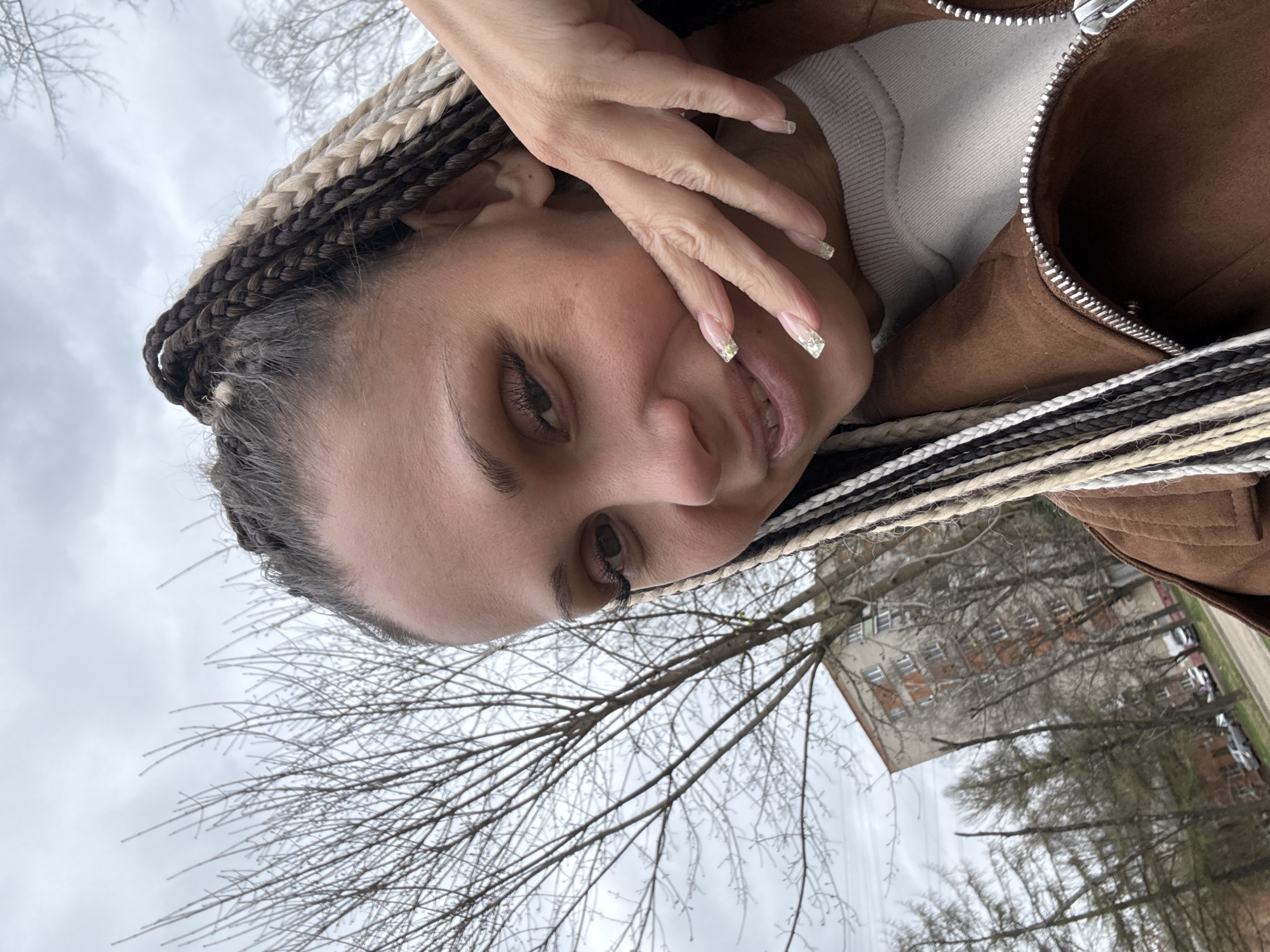 A woman poses outdoors with trees and a building visible in the background. She is wearing a brown jacket and a light-colored shirt underneath. Her hair is styled in braids with alternating brown, black, and white sections. She has manicured nails with a silver or white accent. Her hand is near her face. The building in the background has multiple windows and appears to be located in a residential area. The trees are bare, suggesting it is a cold time of year. It is not possible to confirm the location as Rusanovo, Russia from the available image.