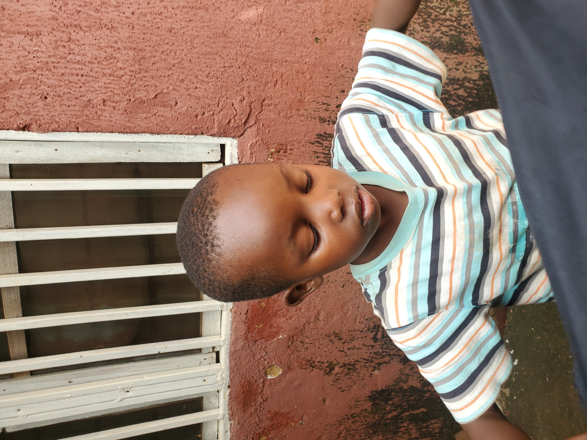 A young Black boy with closed eyes and his head slightly tilted down is positioned in the foreground of the image. He is wearing a horizontally striped t-shirt with alternating bands of light blue, white, gray, and orange. His arms are at his sides, and a dark blue fabric is draped across the lower portion of the frame. The boy is leaning against a reddish-brown, textured wall. To the left of the boy and the wall, there is a window with white-painted bars. The background suggests an outdoor urban environment, potentially in Jos, Nigeria, characterized by the weathered wall and the barred window, common architectural features in the city. The lighting is even, indicating it might be daytime.