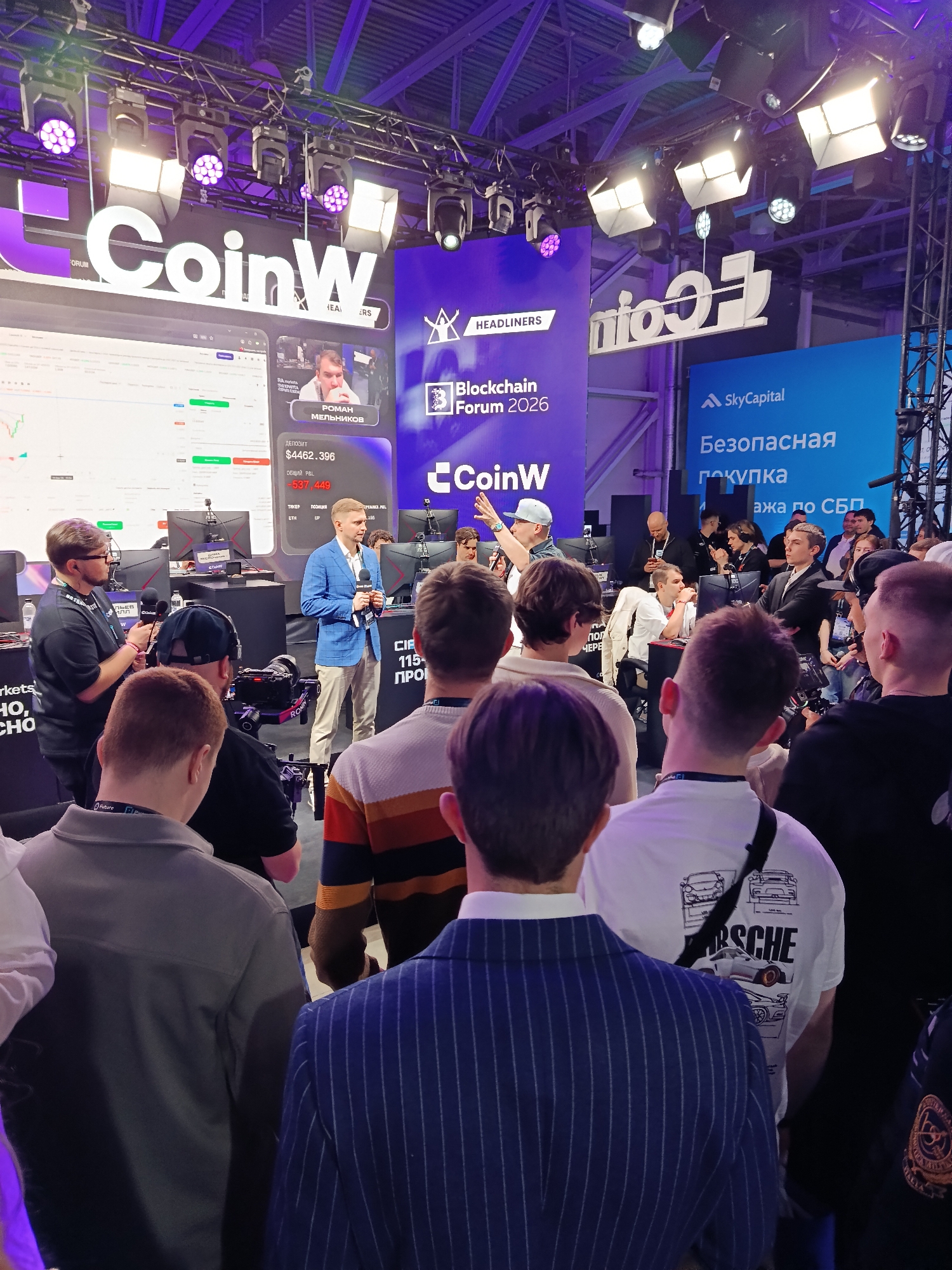 Here is a detailed description of the image:
**Scene:** The image captures an event, likely a conference or presentation, possibly related to blockchain or cryptocurrency, given the presence of "Blockchain Forum 2026" and "CoinW" branding. The location appears to be indoors, with a stage or presentation area at the front. The lighting is a mix of white and purple hues, creating a modern and vibrant atmosphere.
**People:** The image shows a crowd of people, mostly men, facing towards the front of the scene. They are the audience, seemingly listening to a speaker or observing a presentation. One individual in a blue blazer is holding a microphone, suggesting he is speaking or part of a panel. Other people are filming. A man in a white hat gestures with his hand. Individuals are dressed in diverse styles, ranging from casual to formal wear.
**Objects:** Key objects visible include large screens displaying market data and the names of individuals. Signage for "CoinW", "Blockchain Forum 2026", and "SkyCapital" is prominent. Cameras, lighting fixtures, and stage equipment are also visible.
**Actions:** The central action seems to be a presentation or discussion, with an engaged audience. Some audience members are actively listening, while others may be filming or documenting the event.
**Interactions:** The primary interaction is between the speaker/presenter and the audience.
**Environment:** The setting is a modern, well-lit event space with a focus on branding and technology.
**Location:** The city and exact venue are not identifiable in the image.